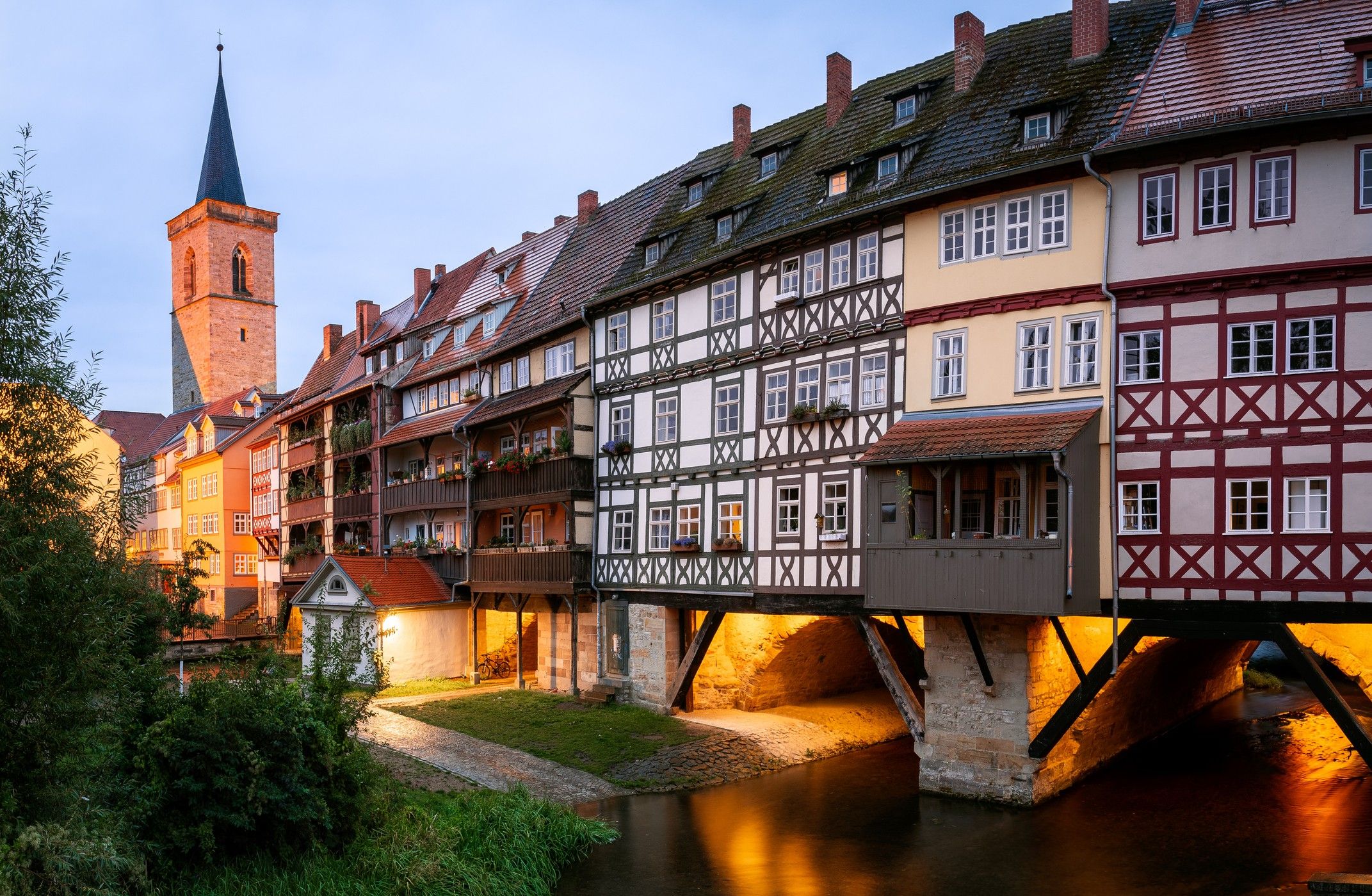 Abendliche Ansicht historischer Fachwerkhäuser, die auf einer Brücke über einen ruhigen Fluss stehen, mit warmem Licht und einem Kirchturm im Hintergrund, vermittelt eine einladende, malerische Atmosphäre einer charmanten Altstadt.