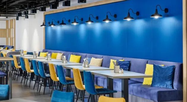 Modernes Restaurant mit langem Tisch, blauen und gelben Stühlen sowie einer blauen Wand mit Wandleuchten. Gemütliche Kissen in Blau und Gelb sorgen für entspannte Atmosphäre.