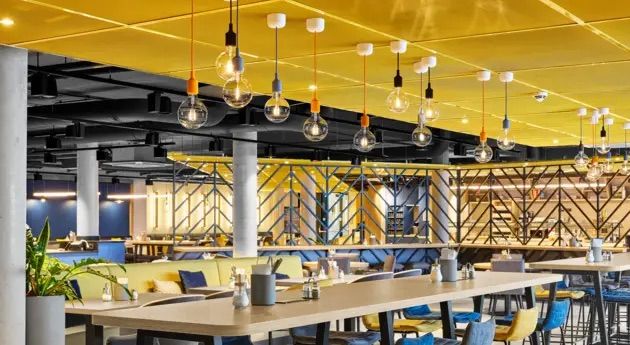 Modernes, helles Restaurant mit großem Gemeinschaftstisch, bunten Stühlen und stilvoller Deckenbeleuchtung für geselliges Essen und entspannte Atmosphäre.