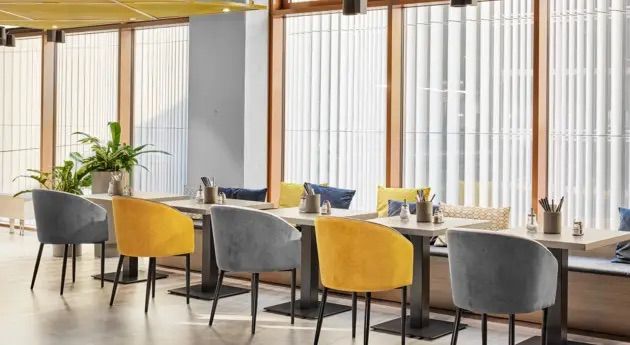 Modernes, helles Restaurant mit großen Fenstern, gemütlichen grauen und gelben Stühlen und sorgfältig gedeckten Tischen. Natürliche Pflanzen sorgen für eine einladende Atmosphäre, ideal für entspannte Mahlzeiten oder Geschäftstreffen.