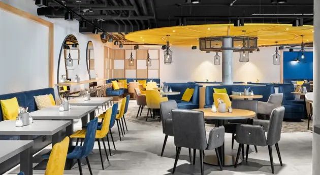 Moderner, heller Hotelrestaurantbereich mit stilvollen blauen und gelben Stühlen, runden und rechteckigen Tischen, gemütlichen Sitzbereichen und warmem, einladendem Ambiente für entspannte Mahlzeiten und geselliges Beisammensein.