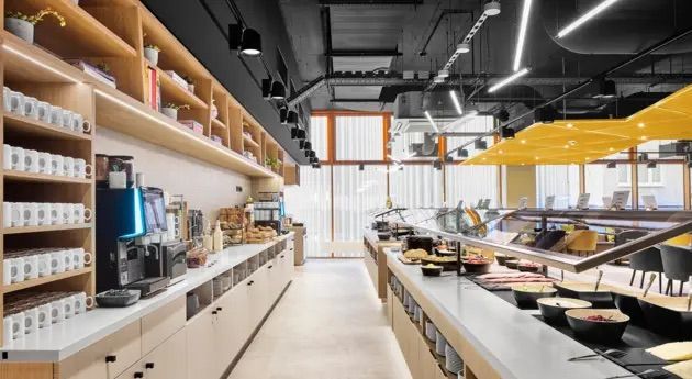 Moderne, helle Hotelbuffetküche mit Frühstücksauswahl, Kaffeemaschine und gemütlichem Sitzbereich. Frische Speisen und Getränke sind ansprechend präsentiert für einen guten Start in den Tag.