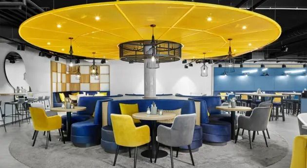Modern gestalteter Restaurantbereich mit rundem blauen Sitzbereich, gelben und grauen Stühlen, gedeckten Tischen und auffälliger großer gelber Deckenleuchte, die eine gemütliche Atmosphäre für entspannte Mahlzeiten schafft.