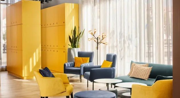 Heller, moderner Lounge-Bereich mit gelben und blauen Sesseln, Sesseln und Sofas, dekorativen Kissen und großen Fenstern, die viel Tageslicht einlassen. Angenehme Atmosphäre für Entspannung und Begegnungen.