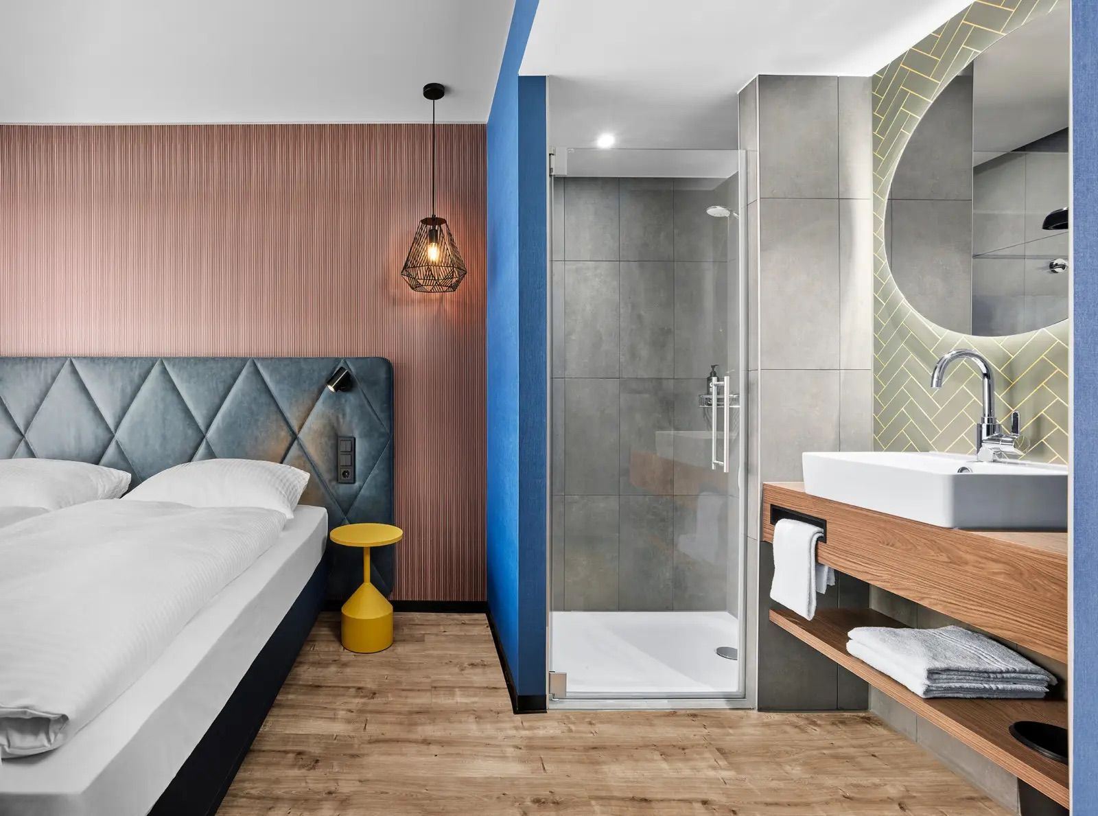 Modernes Hotelzimmer mit gemütlichem Doppelbett, indirekter Beleuchtung und klarem Badbereich mit Dusche, großem Spiegel und praktischem Waschtisch. Helle, warme Farben schaffen eine einladende Atmosphäre für erholsame Nächte und komfortable Aufenthalte.