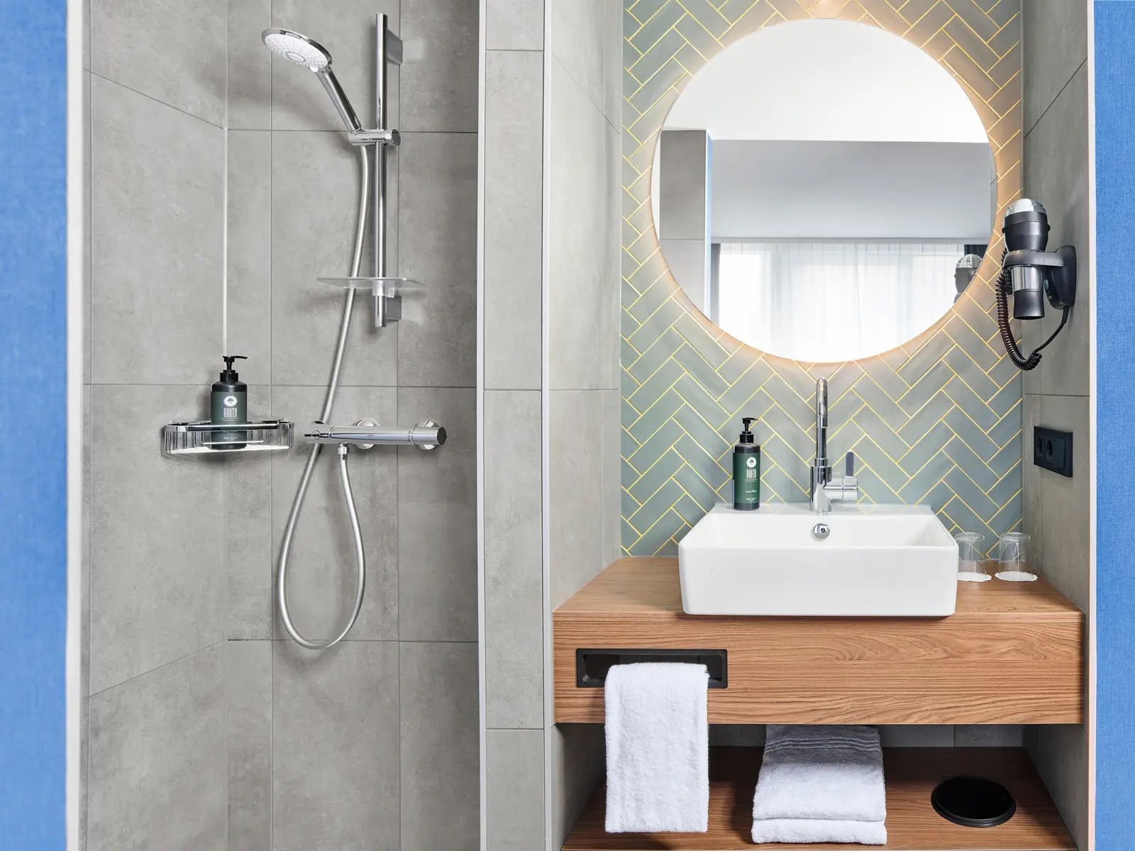Modernes Badezimmer mit grauer Fliesendusche, rechteckigem Waschbecken auf Holzablage, beleuchtetem Rundspiegel und Haartrockner an der Wand. Frische, klare Atmosphäre für Komfort und Entspannung.