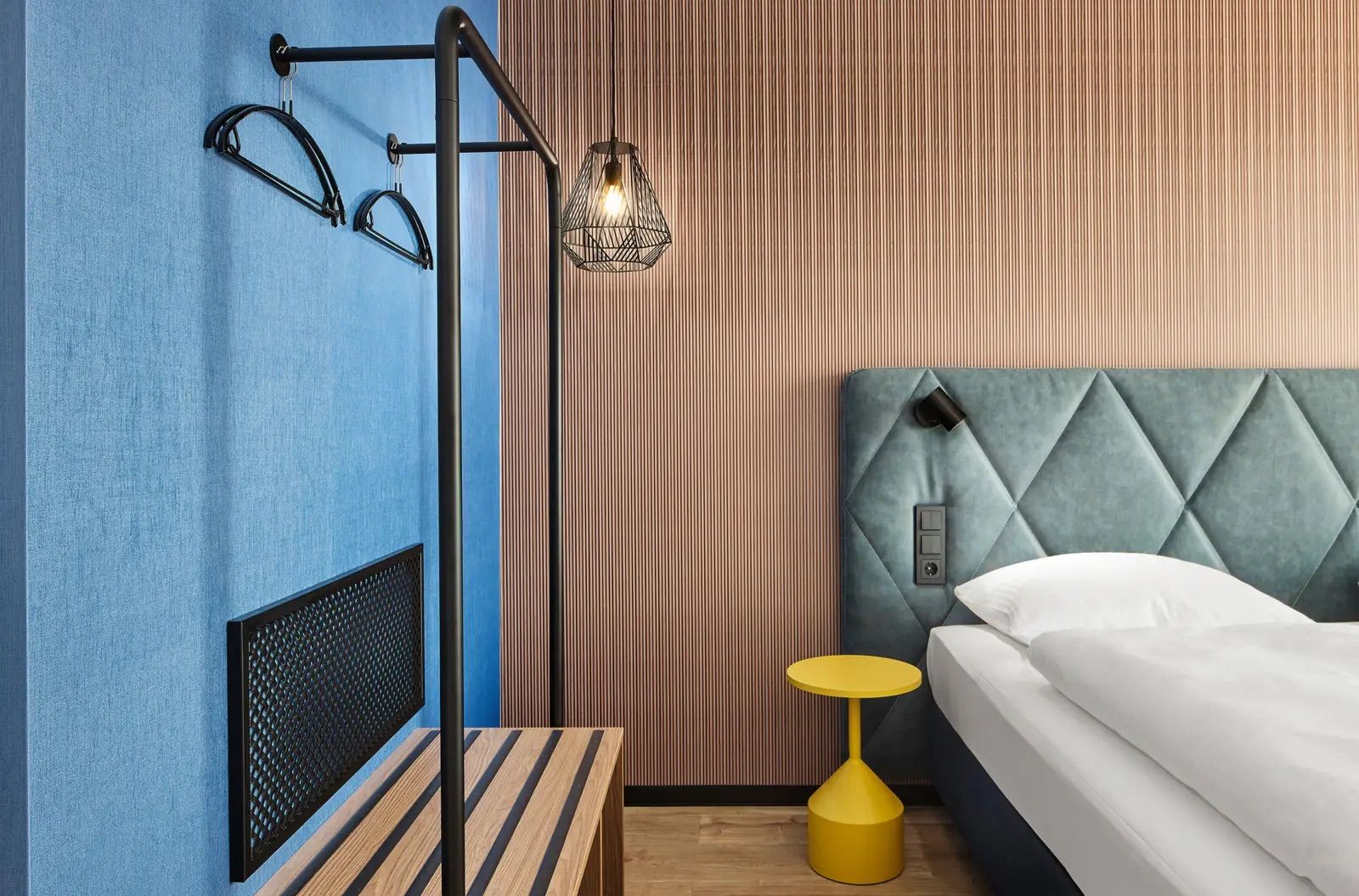Modernes Hotelzimmer mit einem gepolsterten Doppelbett, einem kleinen gelben Nachttisch und einem offenen Kleiderständer aus Metall vor einer blauen Wand, das eine helle, einladende Atmosphäre schafft.