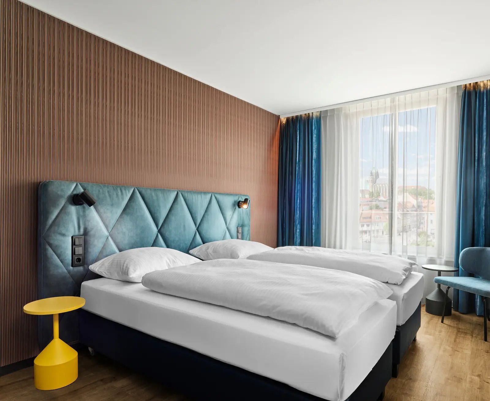Modernes Hotelzimmer mit zwei Einzelbetten, weichen weißen Bettwaren, gepolstertem blauen Kopfteil und großem Fenster mit Blick auf die Stadt. Helle, freundliche Atmosphäre, kombiniert mit stilvollen, farblichen Akzenten und natürlichem Tageslicht.