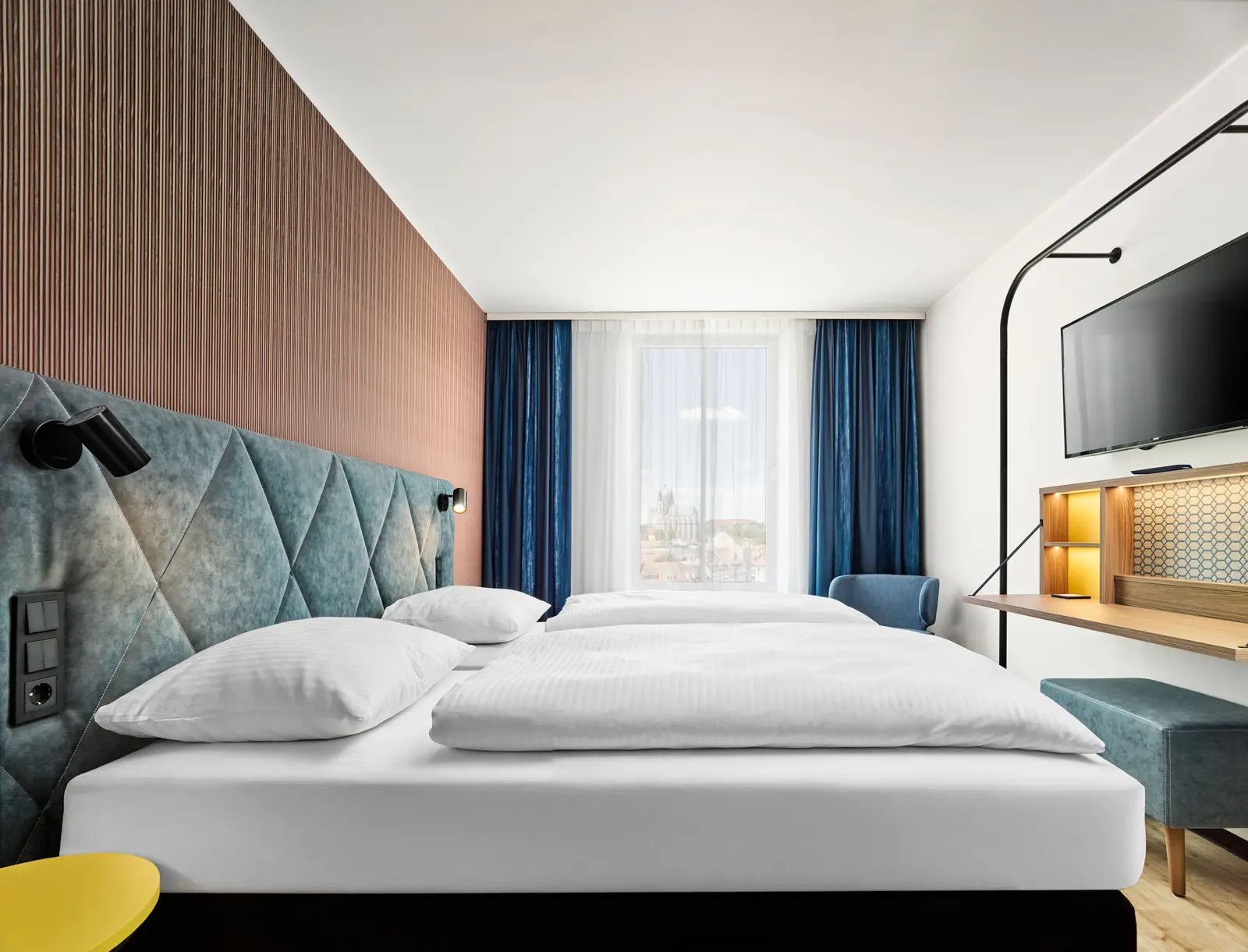 Modernes Hotelzimmer mit großem, bequemem Doppelbett, hellen Vorhängen, stilvoller Holzverkleidung und gemütlichem Sitzbereich. Ruhige, einladende Atmosphäre für erholsamen Aufenthalt.