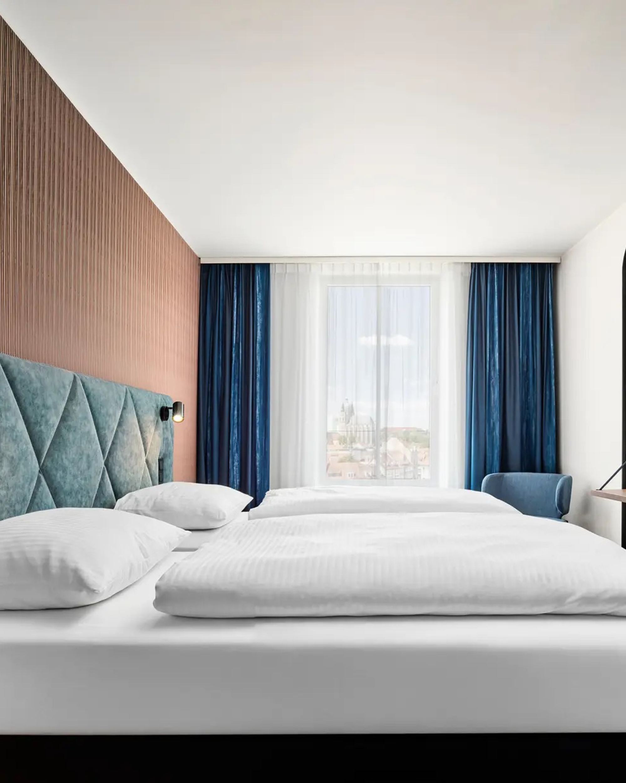 Modernes Hotelzimmer mit großem, bequemem Doppelbett, hellen Vorhängen, stilvoller Holzverkleidung und gemütlichem Sitzbereich. Ruhige, einladende Atmosphäre für erholsamen Aufenthalt.