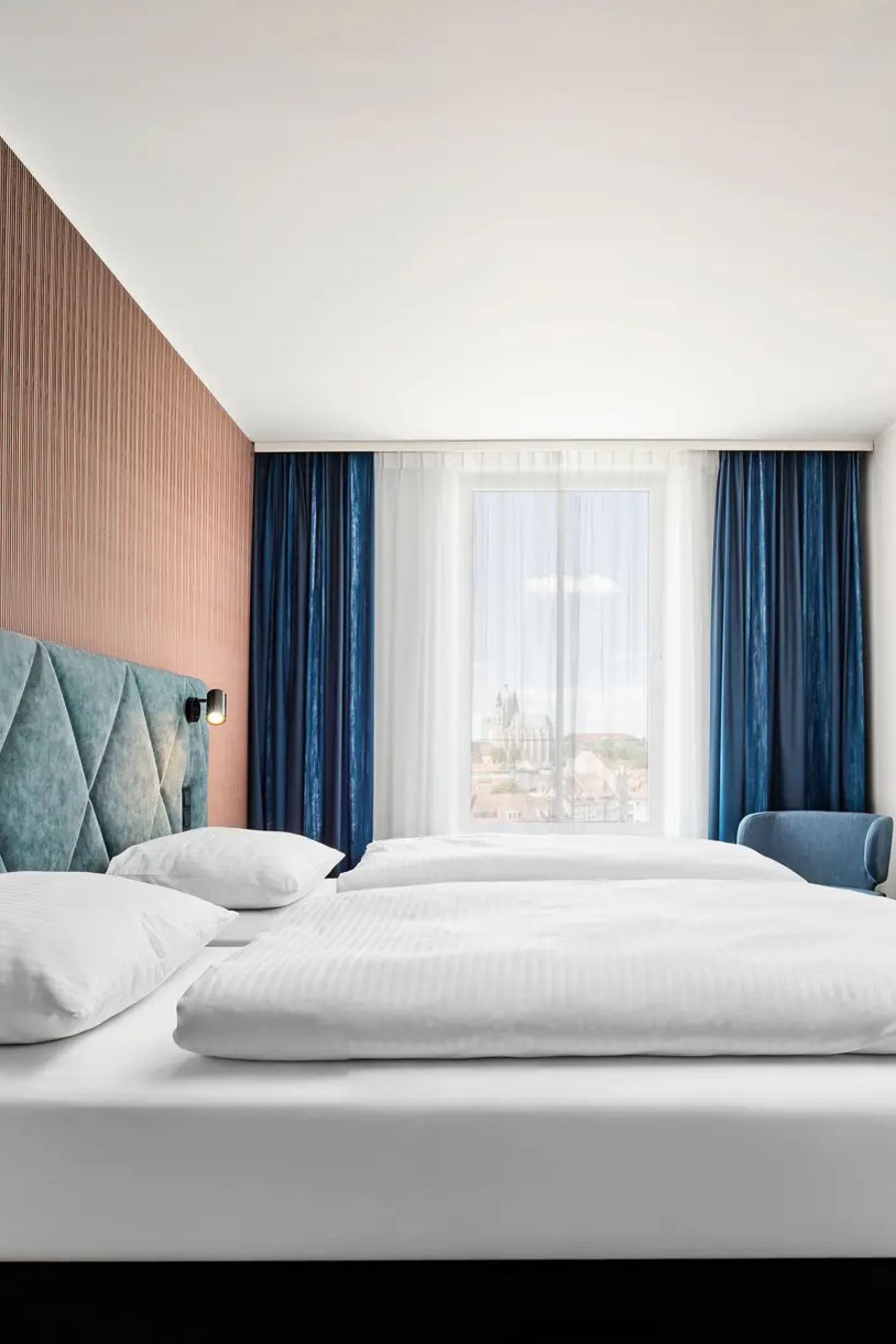 Modernes Hotelzimmer mit großem, bequemem Doppelbett, hellen Vorhängen, stilvoller Holzverkleidung und gemütlichem Sitzbereich. Ruhige, einladende Atmosphäre für erholsamen Aufenthalt.