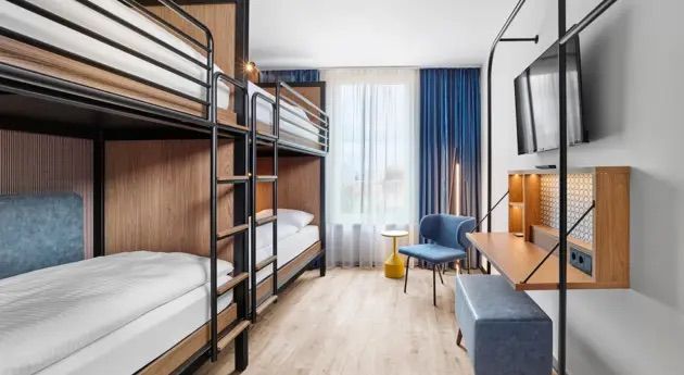 Modernes Zimmer mit Etagenbetten, hellem Holzboden und großen Fenstern. Komfortable Sitzgelegenheit und Arbeitsbereich mit Fernseher schaffen eine praktische, angenehme Atmosphäre für Familien oder Gruppenreisende.