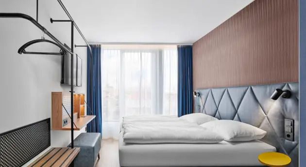 Hotel Erfurt-City Modernes Hotelzimmer mit großem Bett, weichen Kopfkissen, Fenster mit blickdichten Vorhängen und Tageslicht. Helle, freundliche Atmosphäre mit elegantem Holz- und Stoffdesign. Schreibtisch und praktische Ablagen für Reisende.
