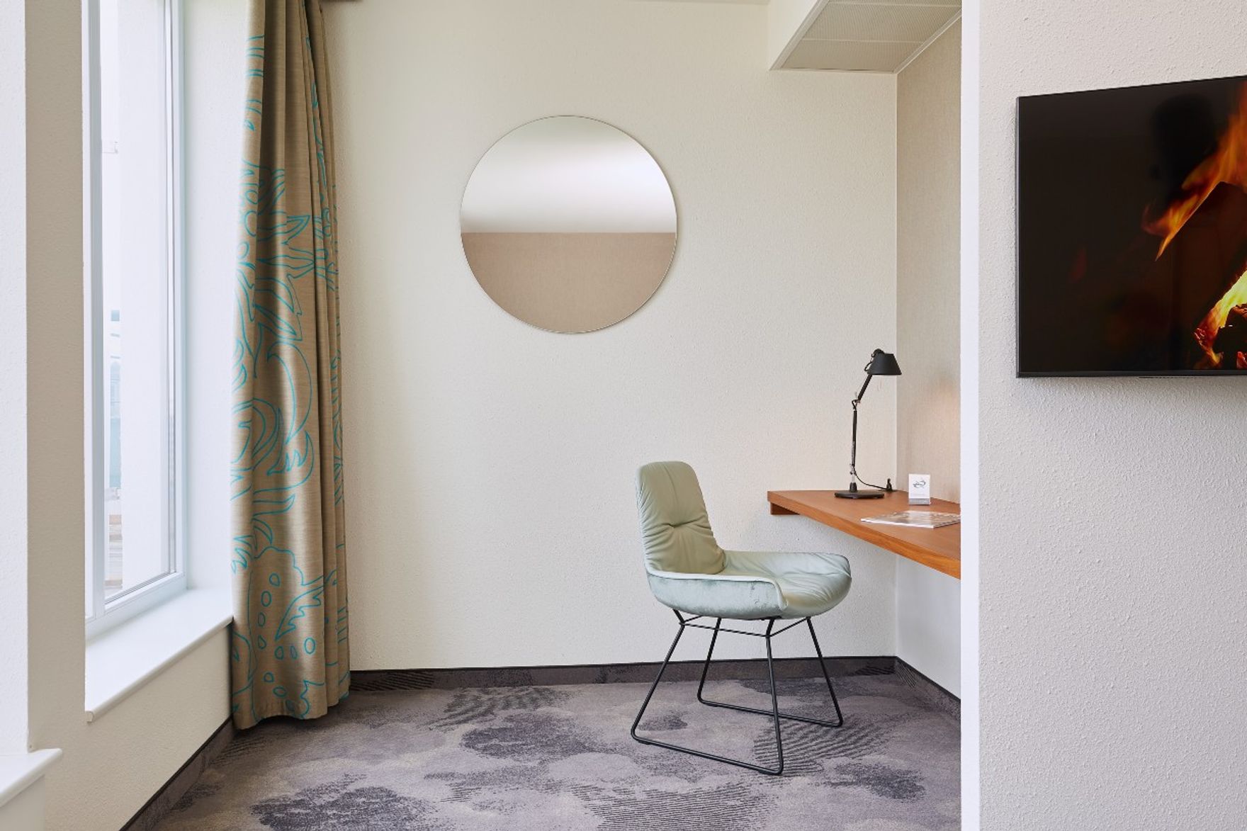 Hotel Essen Motel One lichtdurchflutetes Zimmer mit Blick auf einen Schreibtisch, einen Sessel sowie einen runden Wandspiegel