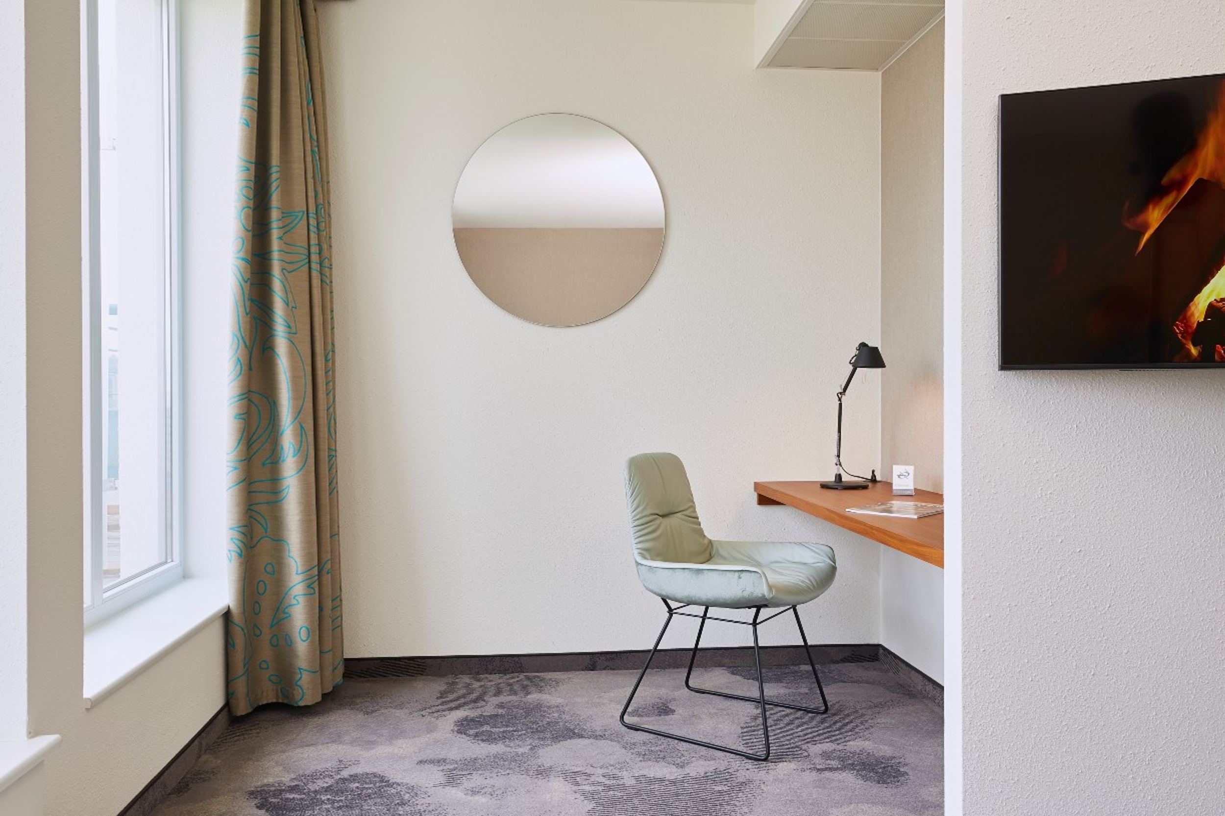 Hotel Essen Motel One lichtdurchflutetes Zimmer mit Blick auf einen Schreibtisch, einen Sessel sowie einen runden Wandspiegel