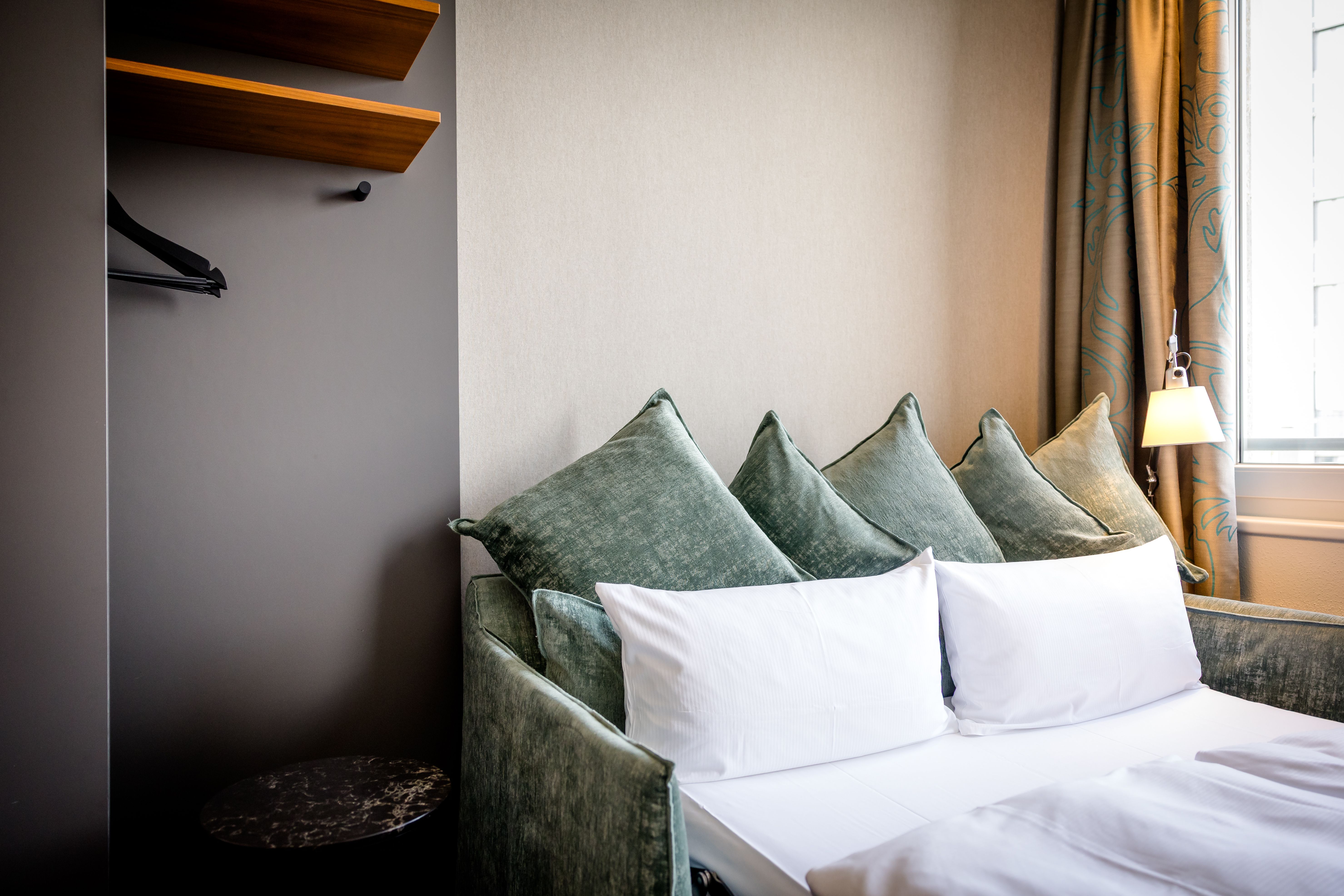 Gemütliches Hotelzimmer mit einem Bett, dekoriert mit weißen Kissen und mehreren grünen Zierkissen, neben einem Fenster mit Vorhang und einer kleinen Wandlampe für angenehmes Licht.