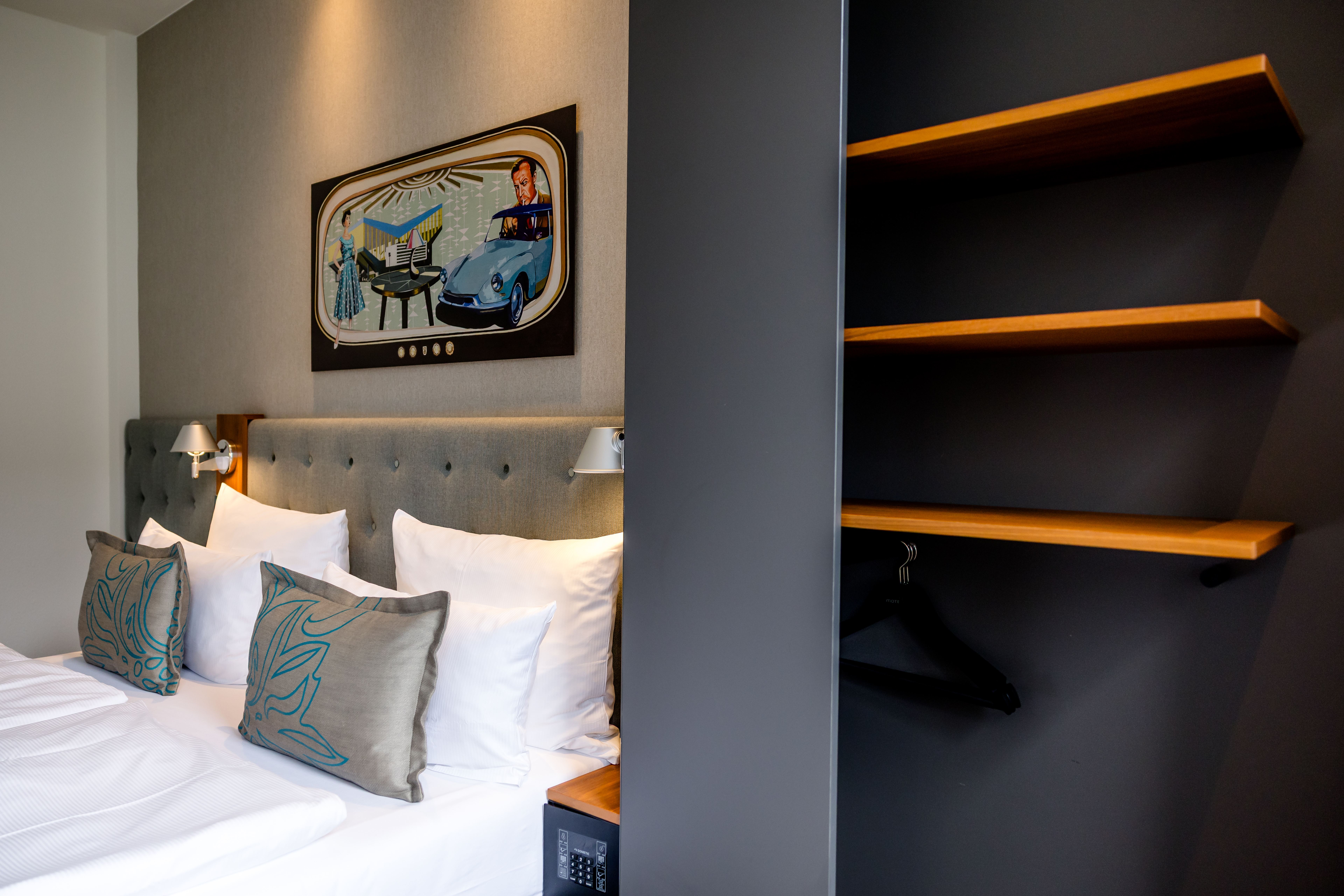 Gemütliches Hotelzimmer mit weißer Bettwäsche, grauem gepolstertem Kopfteil, dekorativen Kissen und moderner Wandbeleuchtung. Nebenan offenliegender Schrank mit Holzregalen und Kleiderbügeln für praktische Aufbewahrung. Ruhige, stilvolle Atmosphäre für erholsamen Aufenthalt. Gemütliches Hotelzimmer mit weißer Bettwäsche, grauem gepolstertem Kopfteil, dekorativen Kissen und moderner Wandbeleuchtung. Nebenan offenliegender Schrank mit Holzregalen und Kleiderbügeln für praktische Aufbewahrung. Ruhige, stilvolle Atmosphäre für erholsamen Aufenthalt.