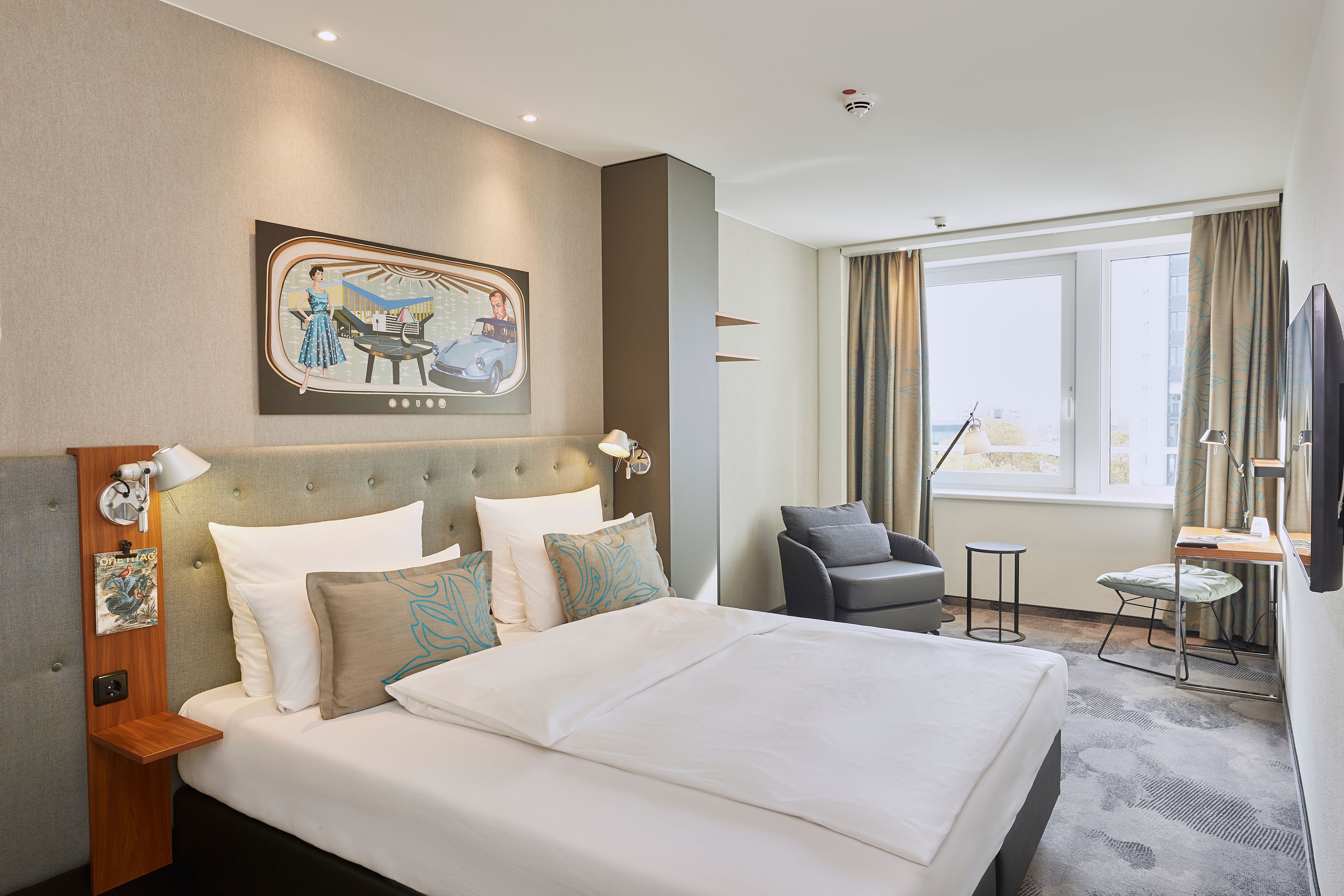 Designhotel Essen Motel One lichtdurchflutetes Zimmer mit geräumigem Doppelbett, Sessel, Schreibtisch und Fernseher an der Wand