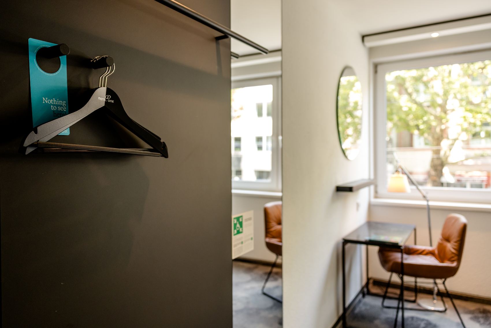 Helle, moderne Hotellounge mit großem Fenster, zwei bequemen Lederstühlen und kleinem Tisch. An der Wand hängen zwei Kleiderbügel, einer mit einem Türanhänger „Nothing to see“. Raum wirkt einladend und funktional für kurze Pausen oder Arbeitszeiten.