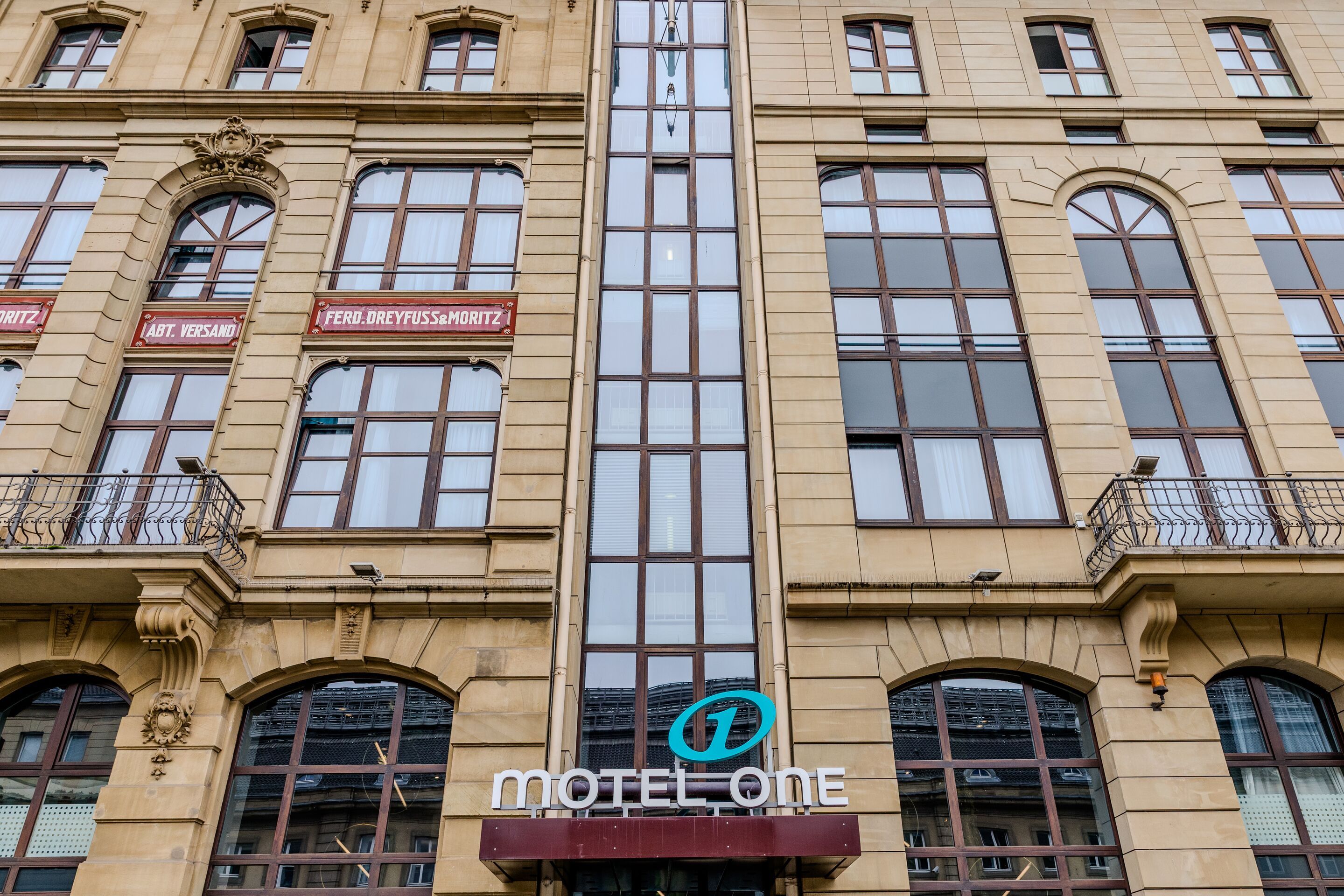 Hotel Frankfurt Hauptbahnhof Motel One Fassade eines Gebäudes mit großen Fenstern, in dessen Mitte ein moderner gläserner Aufzug eingebaut ist, darunter ist das Logo und Schriftzug „Motel One“ zu sehen.