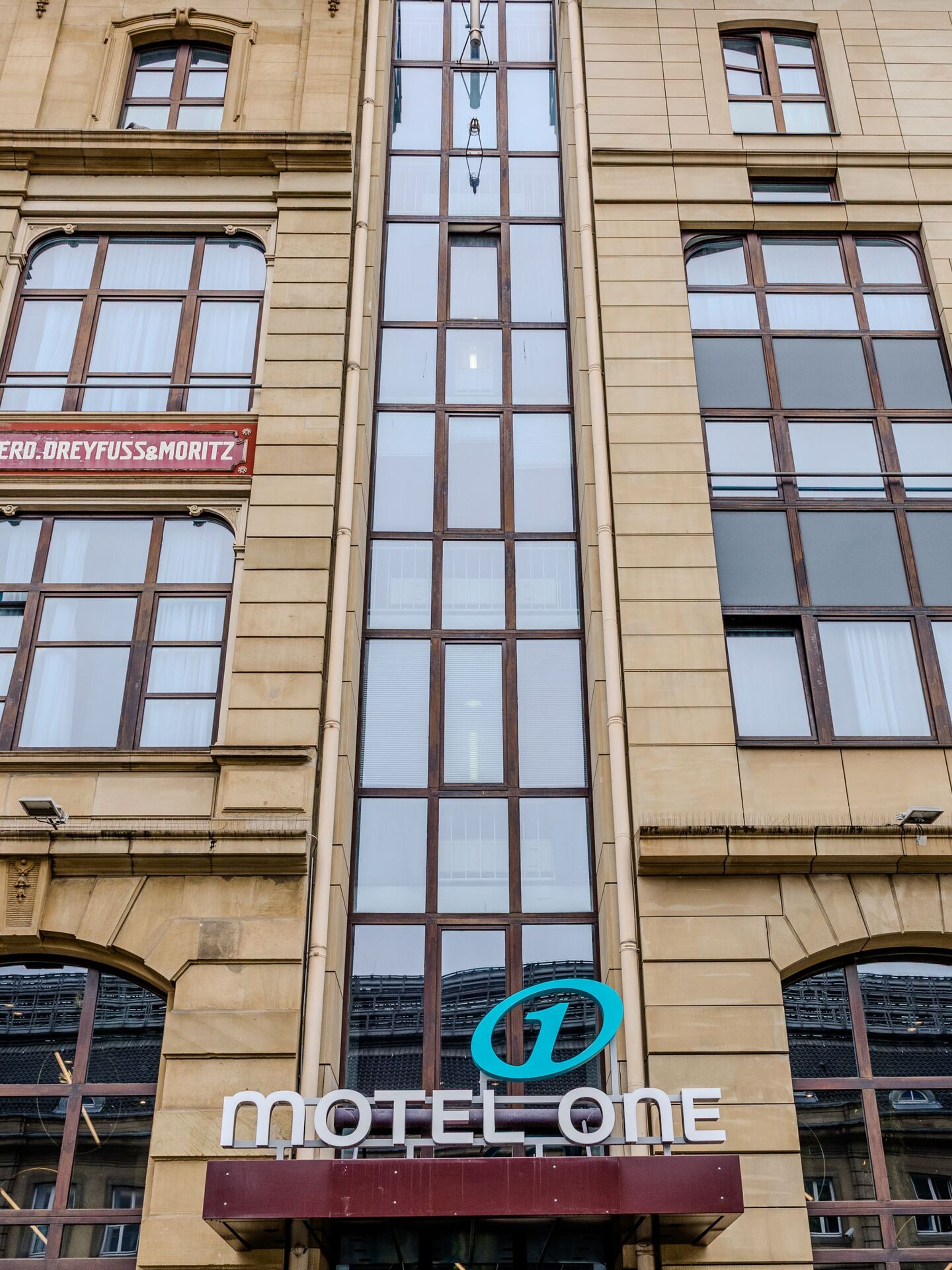 Hotel Frankfurt Hauptbahnhof Motel One Fassade eines Gebäudes mit großen Fenstern, in dessen Mitte ein moderner gläserner Aufzug eingebaut ist, darunter ist das Logo und Schriftzug „Motel One“ zu sehen.