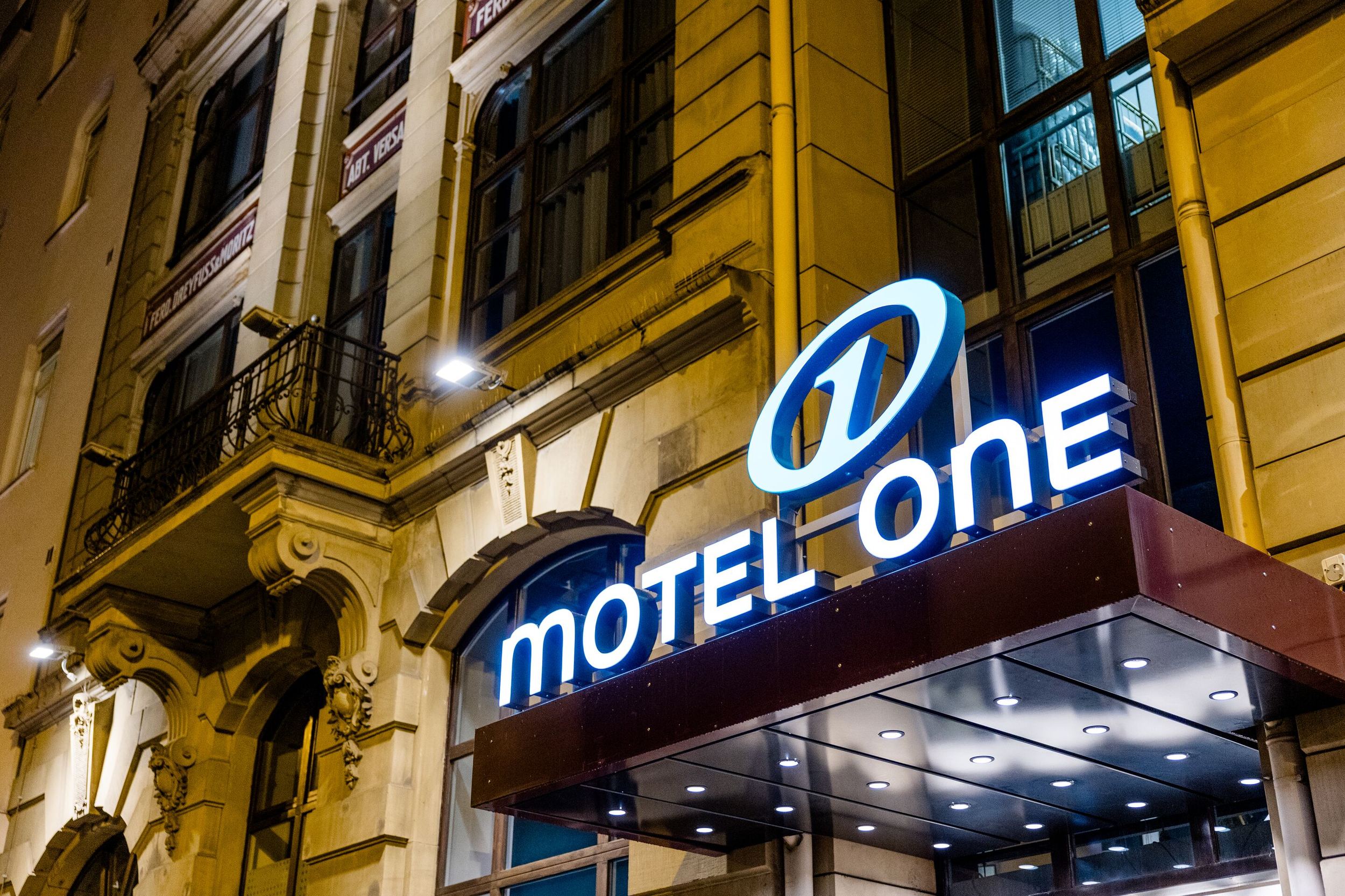 Hotel Frankfurt Hauptbahnhof Motel One beleuchtete Fassade eines historischen Gebäudes mit dem hell erleuchteten Schriftzug „Motel One“ über dem Eingang, der den Hotelstandort in einer belebten Stadtumgebung kennzeichnet.