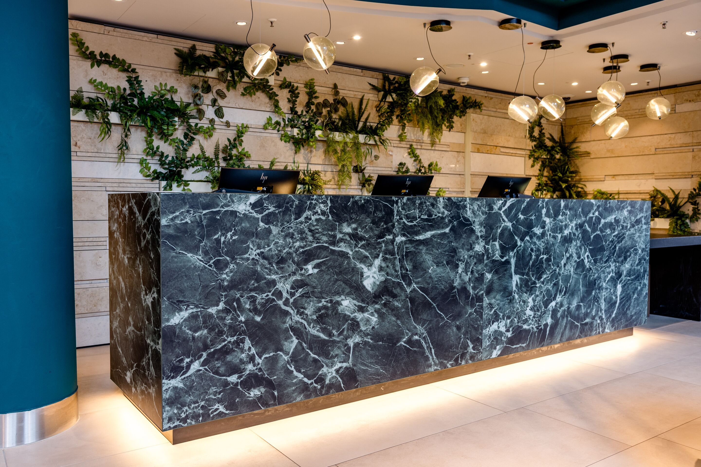 Designhotel Frankfurt-Hauptbahnhof Motel One Rezeption mit Empfangstresen aus dunklem Marmor, warmem Lichtakzent und grünen Pflanzen an der Holzpaneelwand.