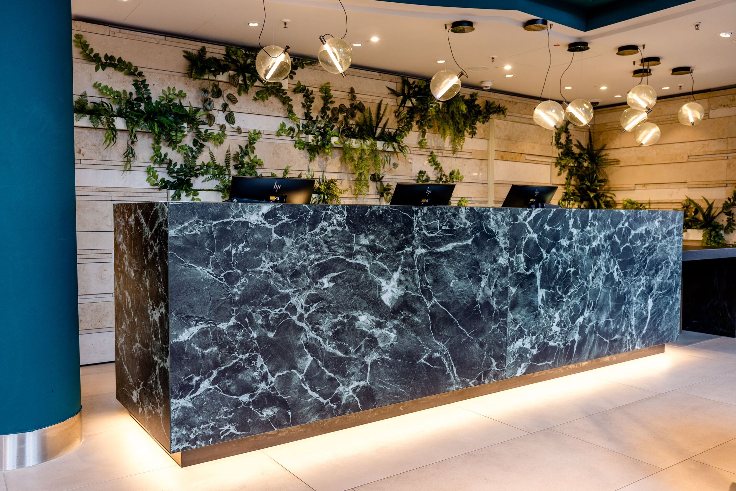 Designhotel Frankfurt-Hauptbahnhof Motel One Rezeption mit Empfangstresen aus dunklem Marmor, warmem Lichtakzent und grünen Pflanzen an der Holzpaneelwand.