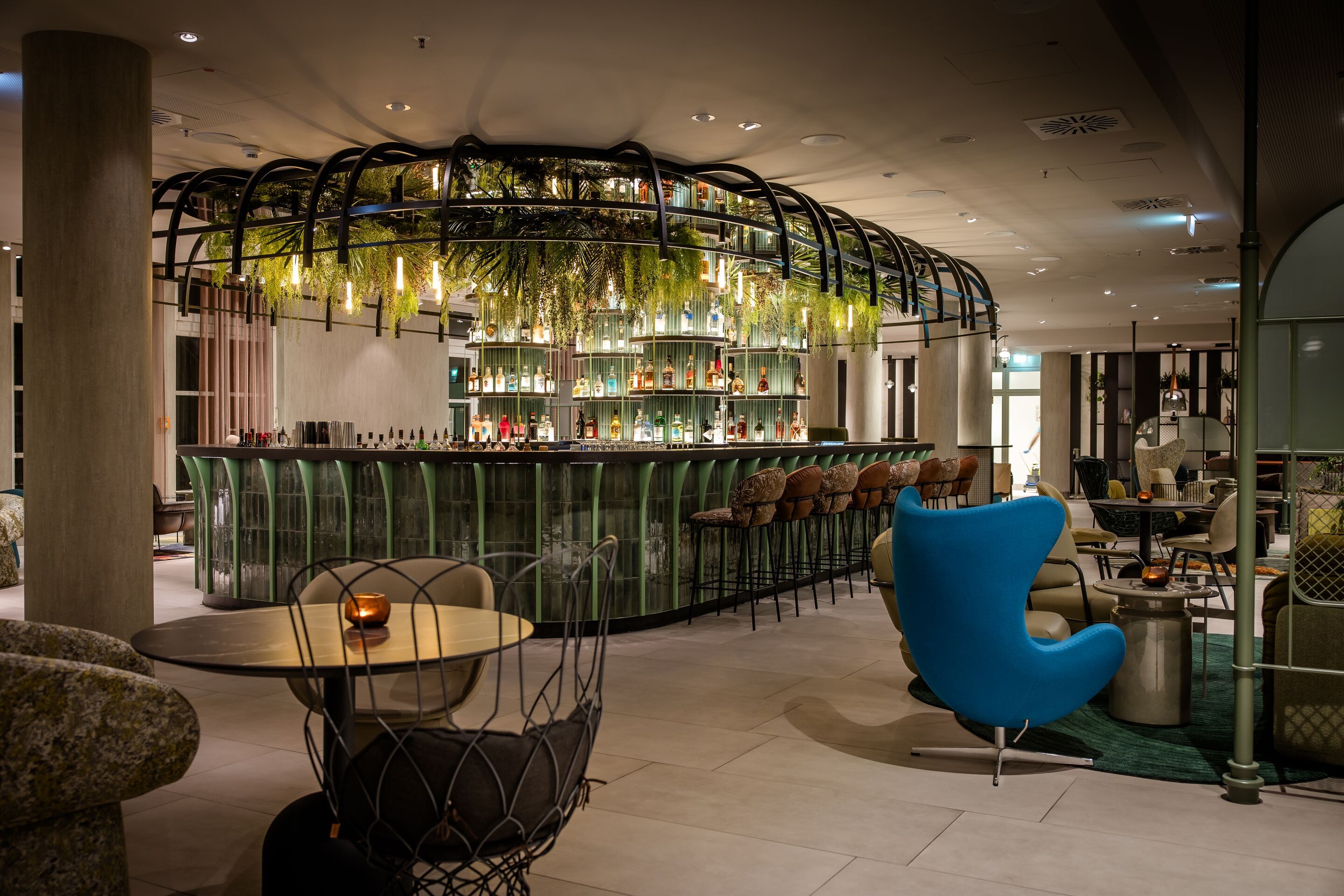 Hotel Frankfurt-Hauptbahnhof Motel One Bar mit grünem Tresen, hängenden Pflanzen und warmem Licht, umgeben von gemütlichen Sitzbereichen mit Lounge-Stühlen und Tischen für entspannte Abende.