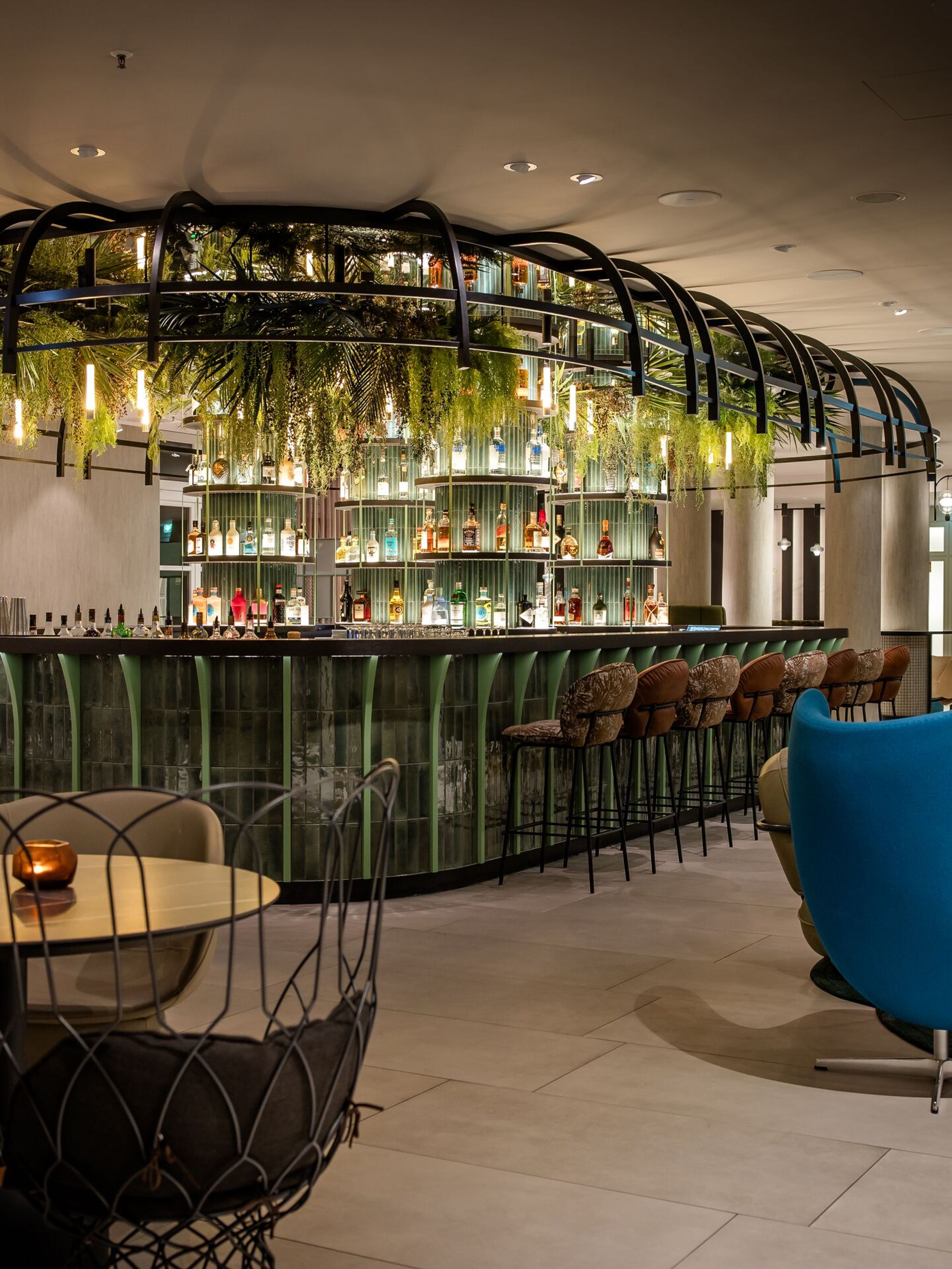 Hotel Frankfurt-Hauptbahnhof Motel One Bar mit grünem Tresen, hängenden Pflanzen und warmem Licht, umgeben von gemütlichen Sitzbereichen mit Lounge-Stühlen und Tischen für entspannte Abende.