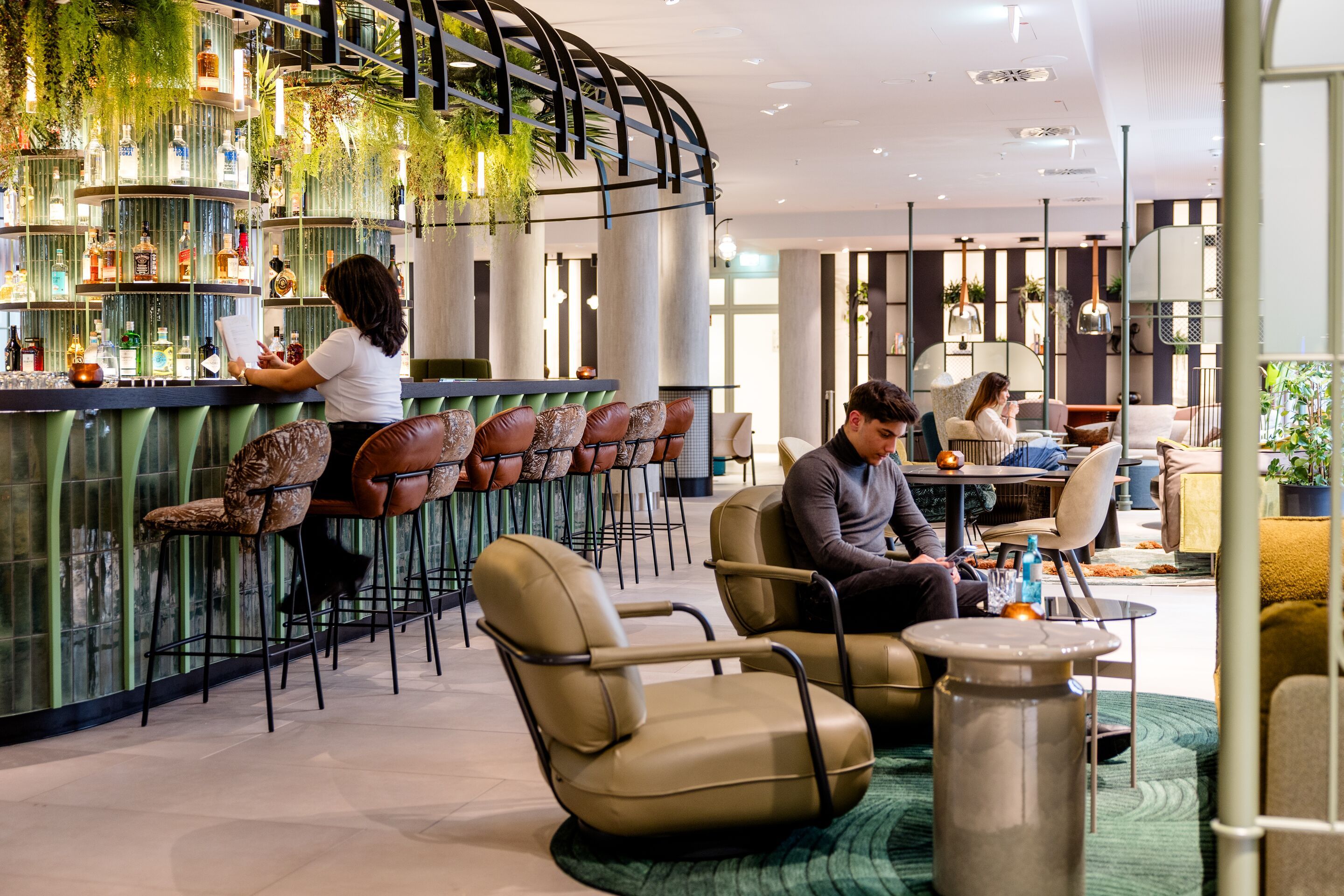 Hotel Frankfurt-Hauptbahnhof Motel One helle Hotelbar mit hängender Pflanzen-Dekoration, bequemen Barhockern und Sitzbereichen. Gäste entspannen beim Lesen und an der Bar und Blick in lichtdurchflutete Lounge.