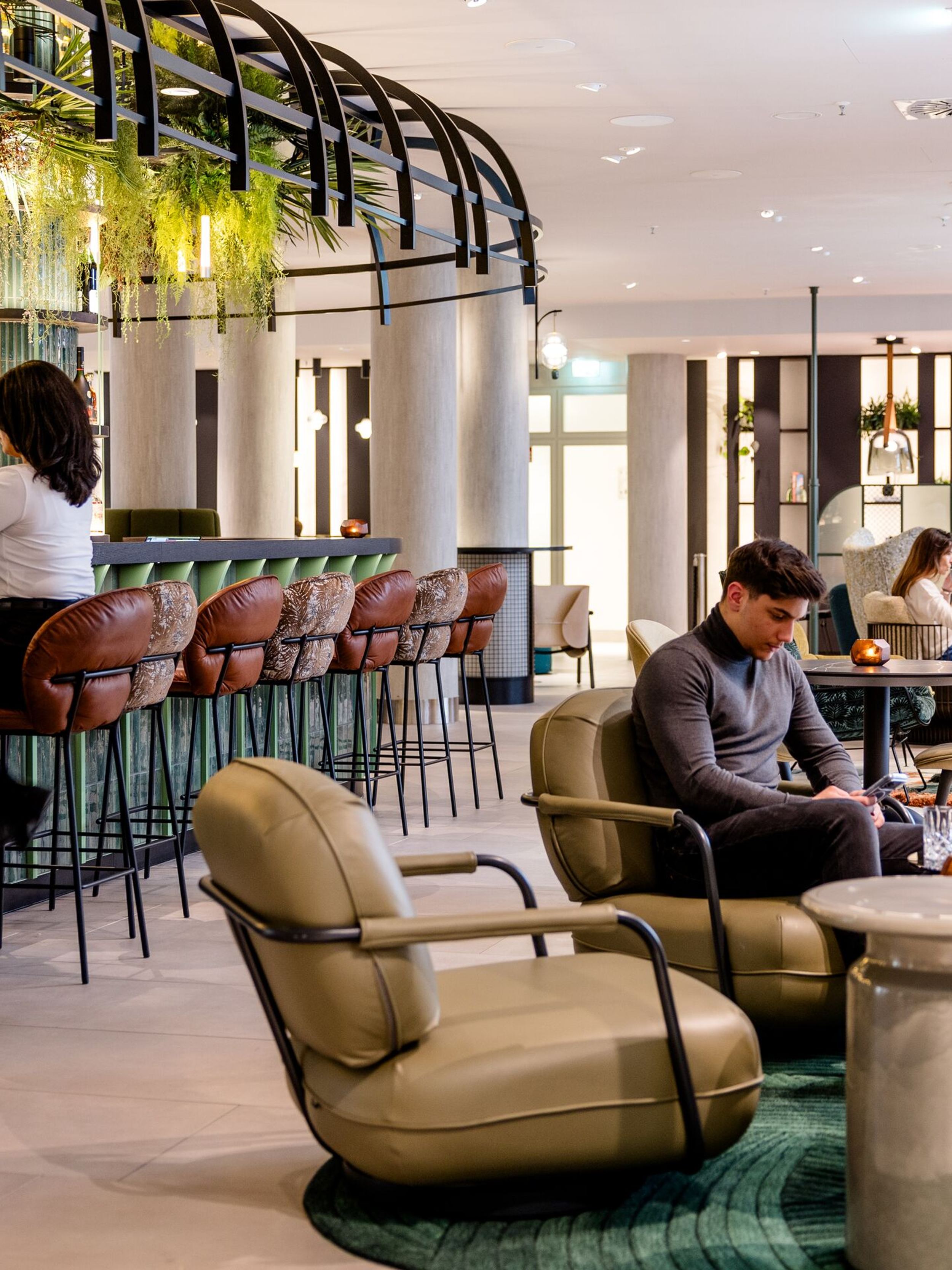 Hotel Frankfurt-Hauptbahnhof Motel One helle Hotelbar mit hängender Pflanzen-Dekoration, bequemen Barhockern und Sitzbereichen. Gäste entspannen beim Lesen und an der Bar und Blick in lichtdurchflutete Lounge.