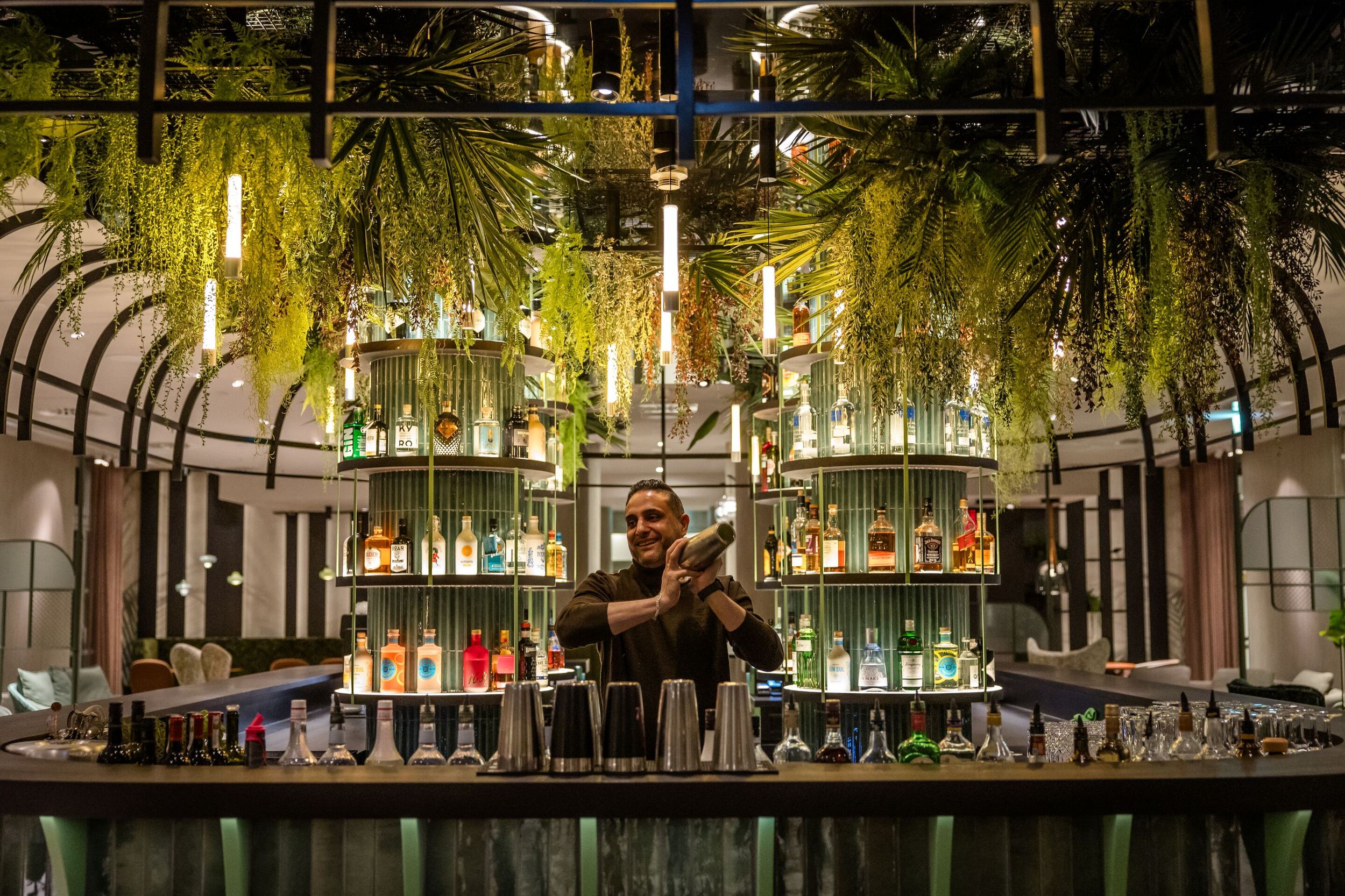 Hotel Frankfurt-Hauptbahnhof Motel One Barkeeper mixt Cocktails an Bar mit hängenden Pflanzen und beleuchteten Regalen voller bunter Spirituosenflaschen in einem eleganten, grünen Ambiente.