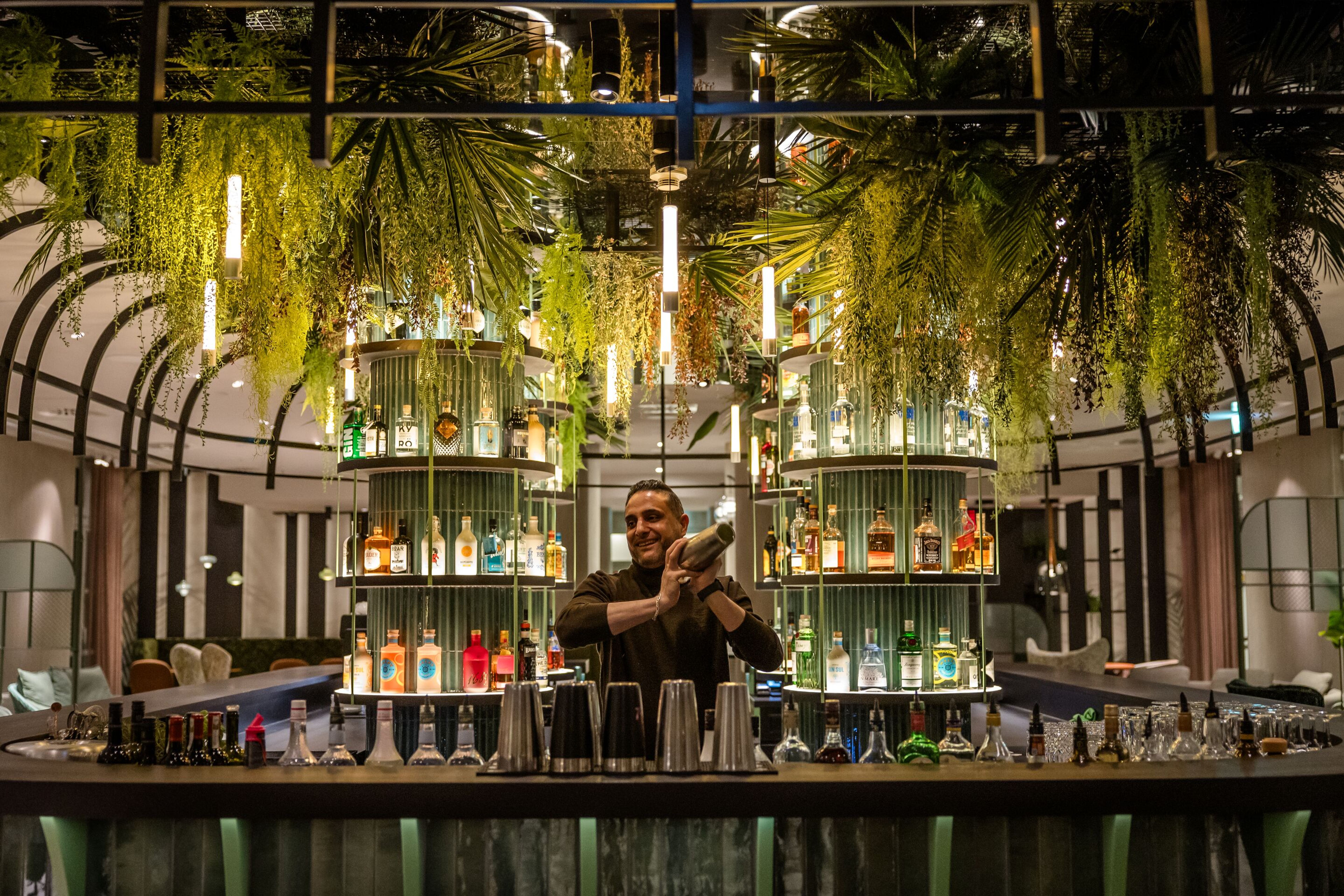 Hotel Frankfurt-Hauptbahnhof Motel One Barkeeper mixt Cocktails an Bar mit hängenden Pflanzen und beleuchteten Regalen voller bunter Spirituosenflaschen in einem eleganten, grünen Ambiente. Hotel Frankfurt-Hauptbahnhof Motel One Barkeeper mixt Cocktails an Bar mit hängenden Pflanzen und beleuchteten Regalen voller bunter Spirituosenflaschen in einem eleganten, grünen Ambiente.