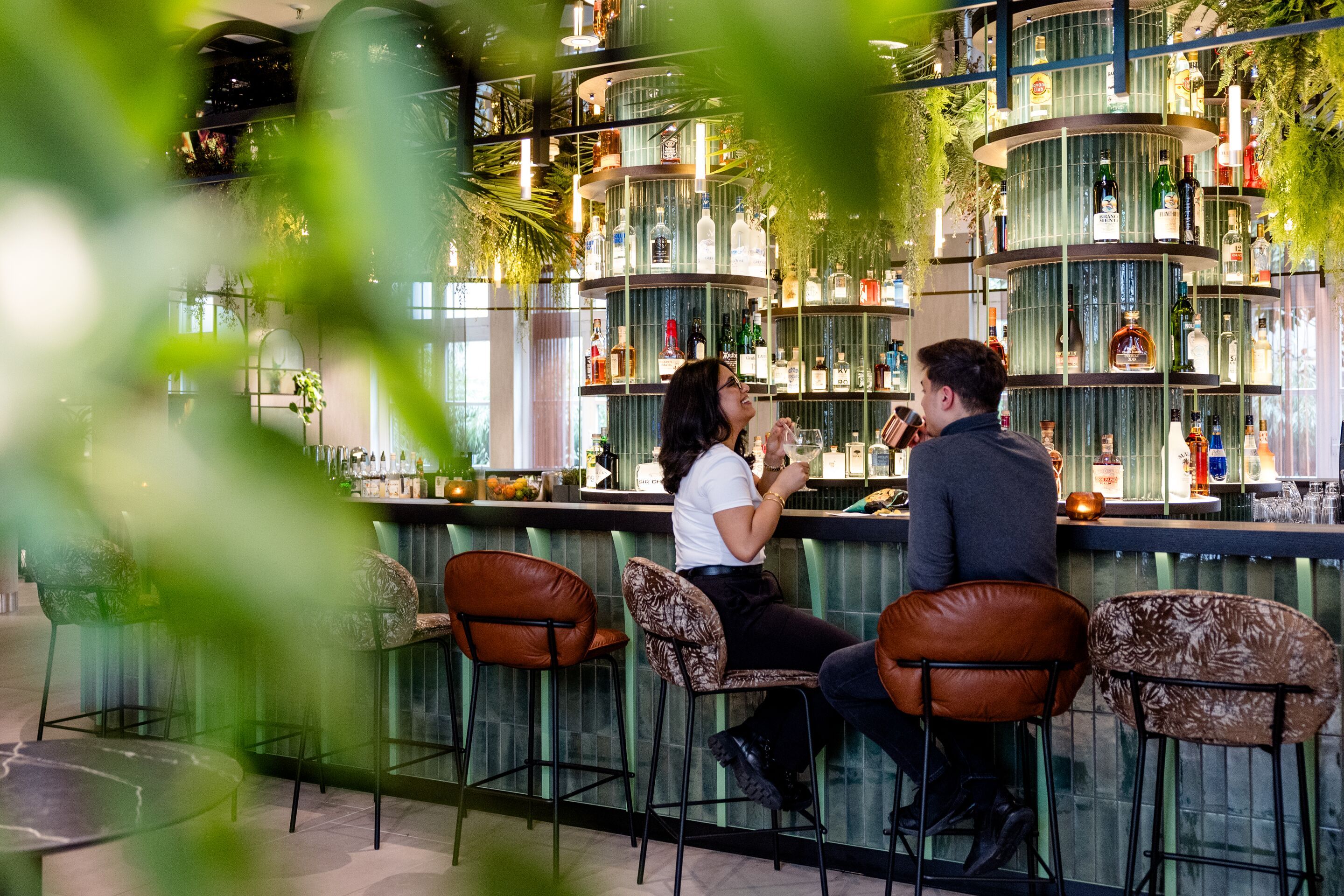 Hotel Frankfurt-Hauptbahnhof Motel One zwei Personen sitzen an einer stilvollen Hotelbar und genießen Getränke. Die Bar ist modern gestaltet mit dekorativen Flaschen und warmem Licht, umgeben von Barhockern und begrüntem Ambiente.