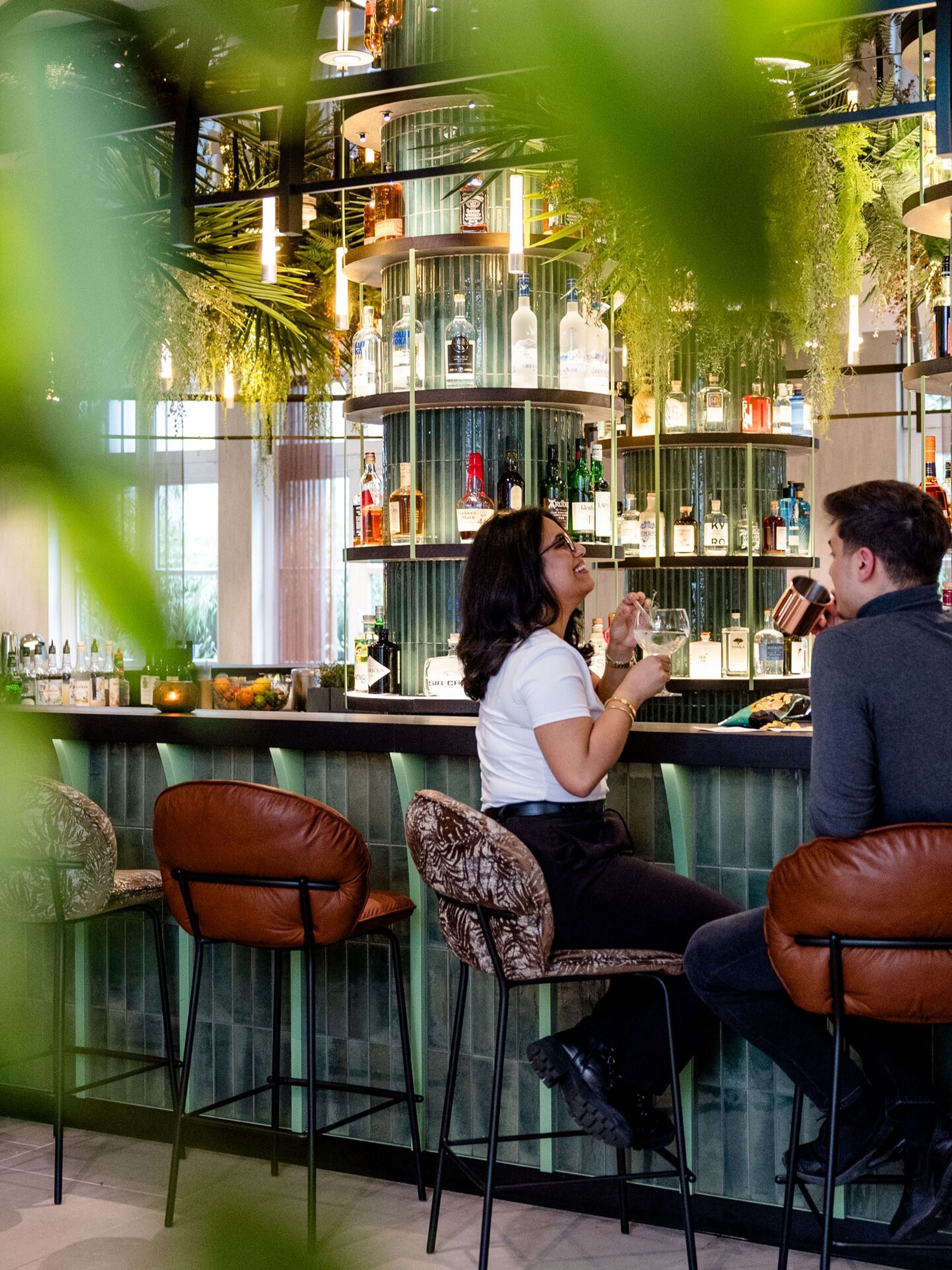 Hotel Frankfurt-Hauptbahnhof Motel One zwei Personen sitzen an einer stilvollen Hotelbar und genießen Getränke. Die Bar ist modern gestaltet mit dekorativen Flaschen und warmem Licht, umgeben von Barhockern und begrüntem Ambiente.