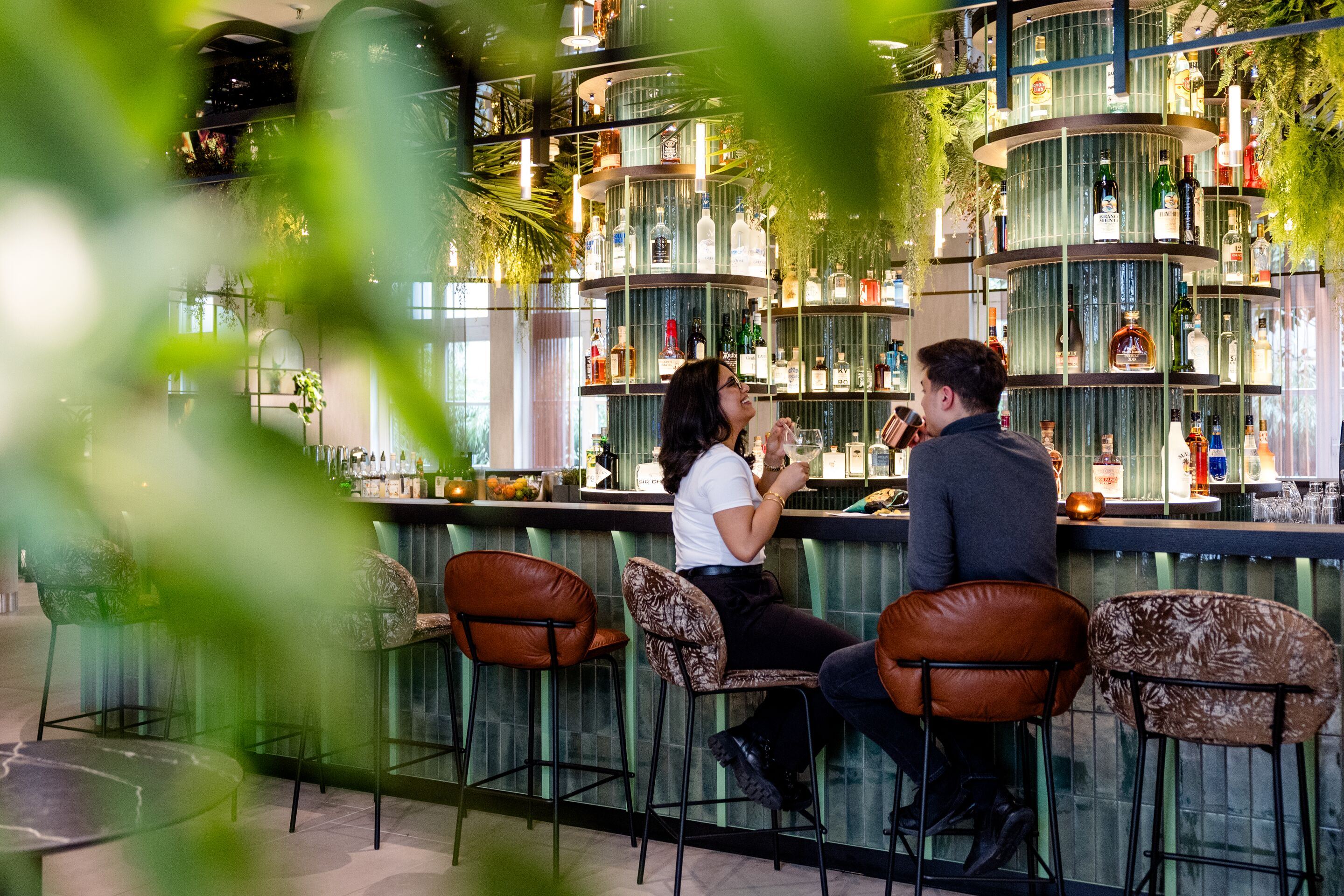 Hotel Frankfurt-Hauptbahnhof Motel One zwei Personen sitzen an einer stilvollen Hotelbar und genießen Getränke. Die Bar ist modern gestaltet mit dekorativen Flaschen und warmem Licht, umgeben von Barhockern und begrüntem Ambiente. Hotel Frankfurt-Hauptbahnhof Motel One zwei Personen sitzen an einer stilvollen Hotelbar und genießen Getränke. Die Bar ist modern gestaltet mit dekorativen Flaschen und warmem Licht, umgeben von Barhockern und begrüntem Ambiente.