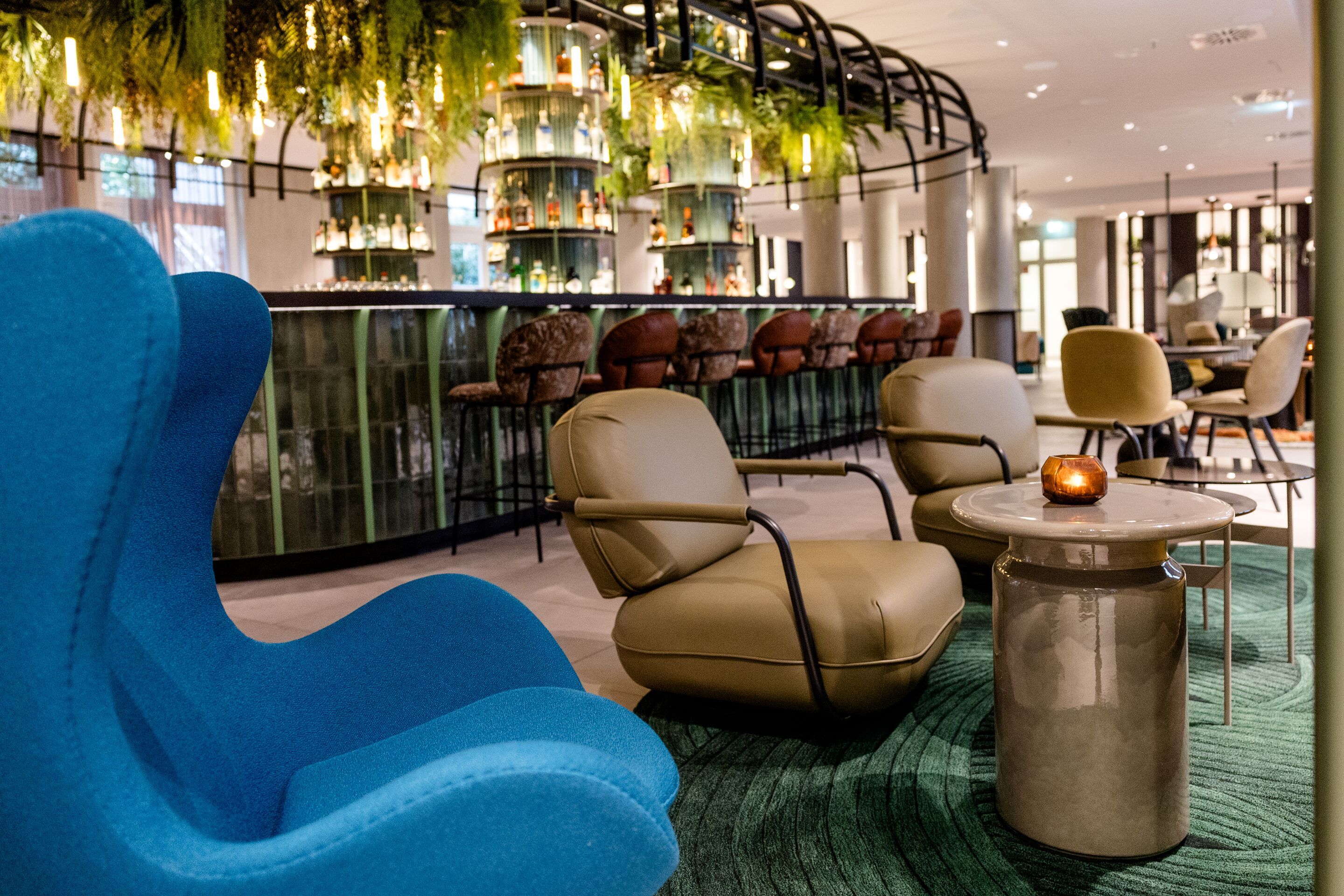 Hotel Frankfurt-Hauptbahnhof Motel One Bar mit Egg Chair und weiteren Stühlen, rundem Tisch mit Kerze, grünem Teppich und hängenden Pflanzen über der Bar.