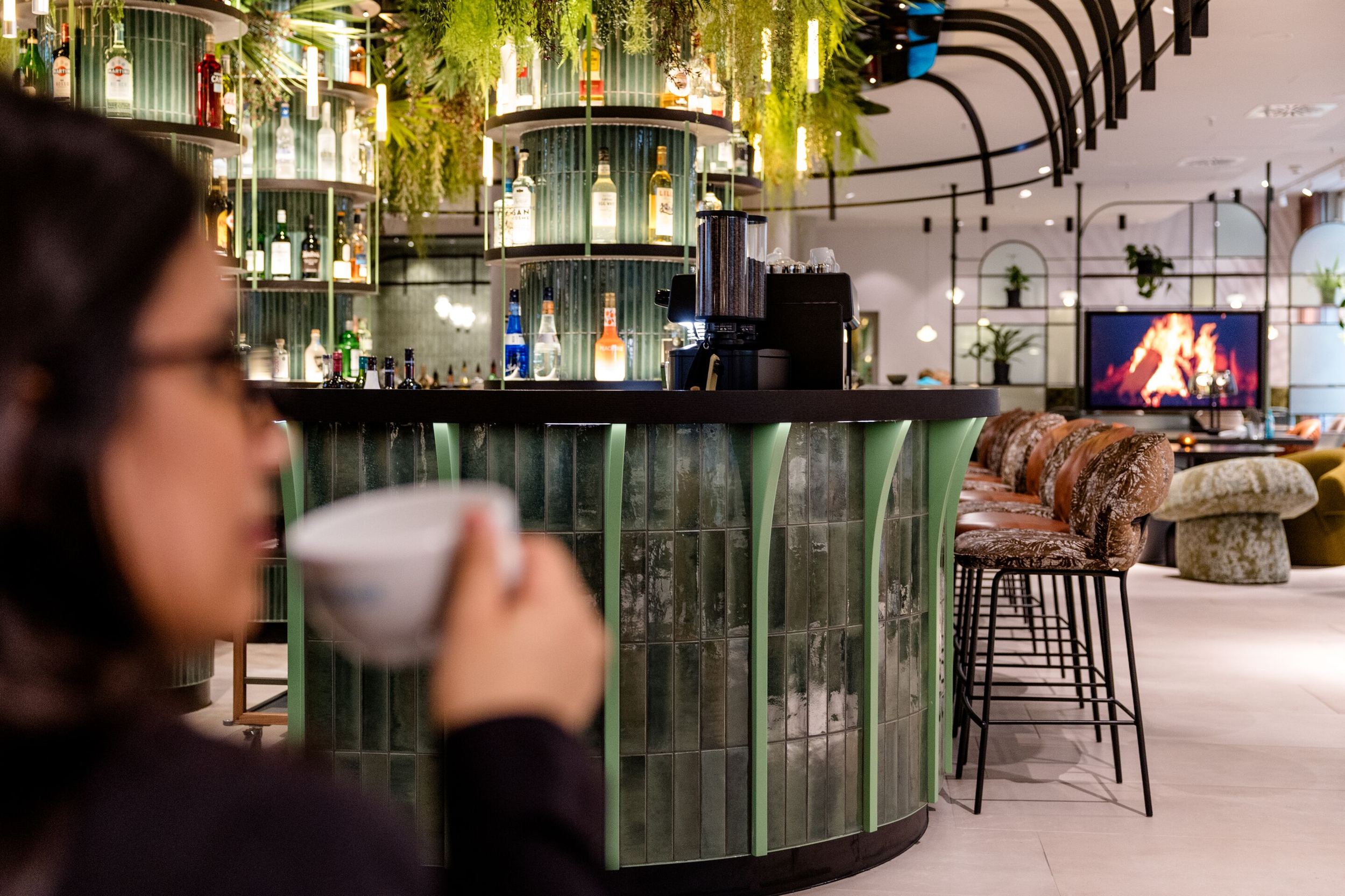 Hotel Frankfurt-Hauptbahnhof Motel One Bar mit grün beleuchtetem Tresen, Barhockern und gemütlichem Lounge-Bereich vor einem digitalen Kaminfeuer, ideal für entspannte Abende und gesellige Momente.