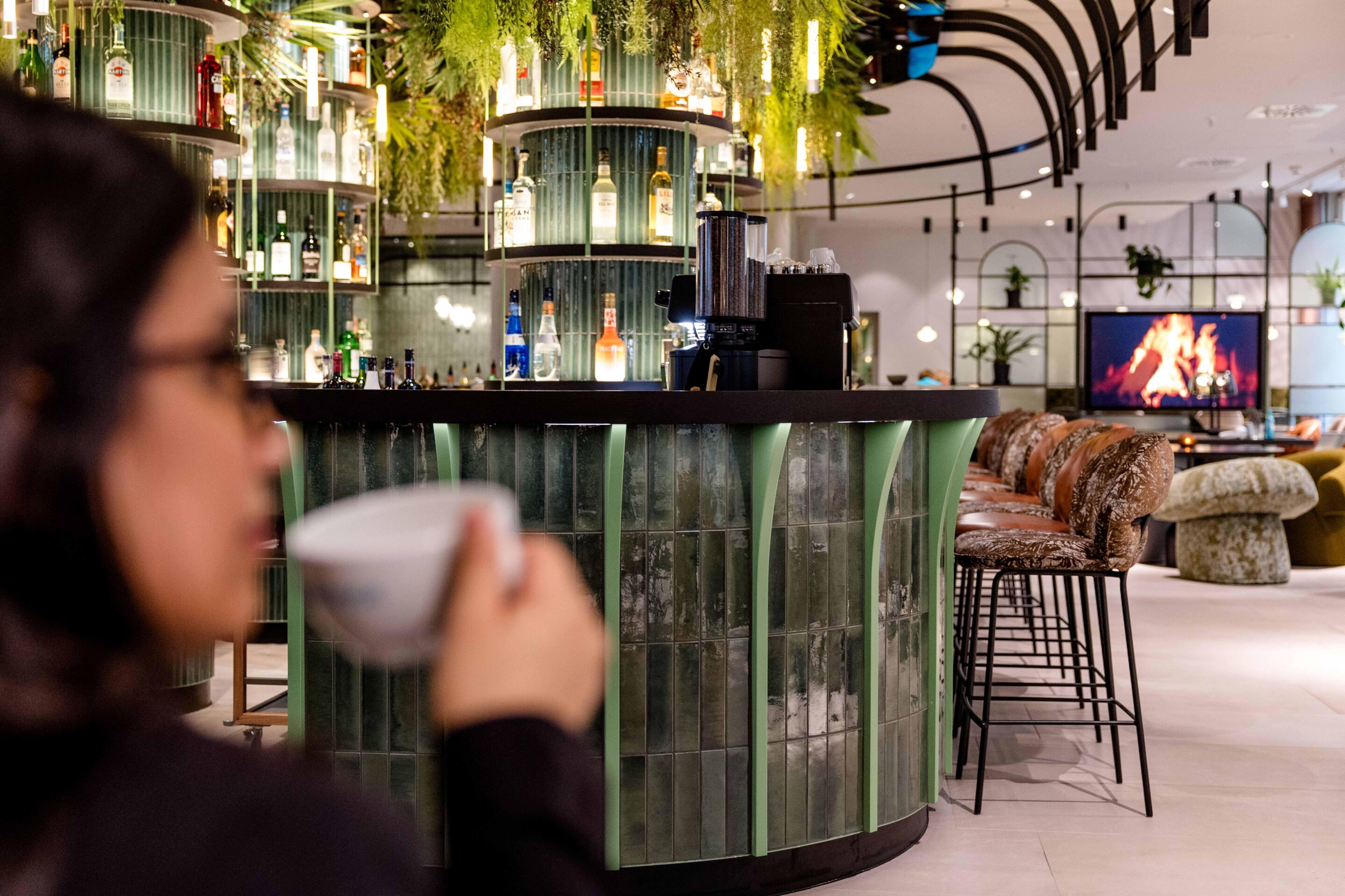 Hotel Frankfurt-Hauptbahnhof Motel One Bar mit grün beleuchtetem Tresen, Barhockern und gemütlichem Lounge-Bereich vor einem digitalen Kaminfeuer, ideal für entspannte Abende und gesellige Momente. Hotel Frankfurt-Hauptbahnhof Motel One Bar mit grün beleuchtetem Tresen, Barhockern und gemütlichem Lounge-Bereich vor einem digitalen Kaminfeuer, ideal für entspannte Abende und gesellige Momente.