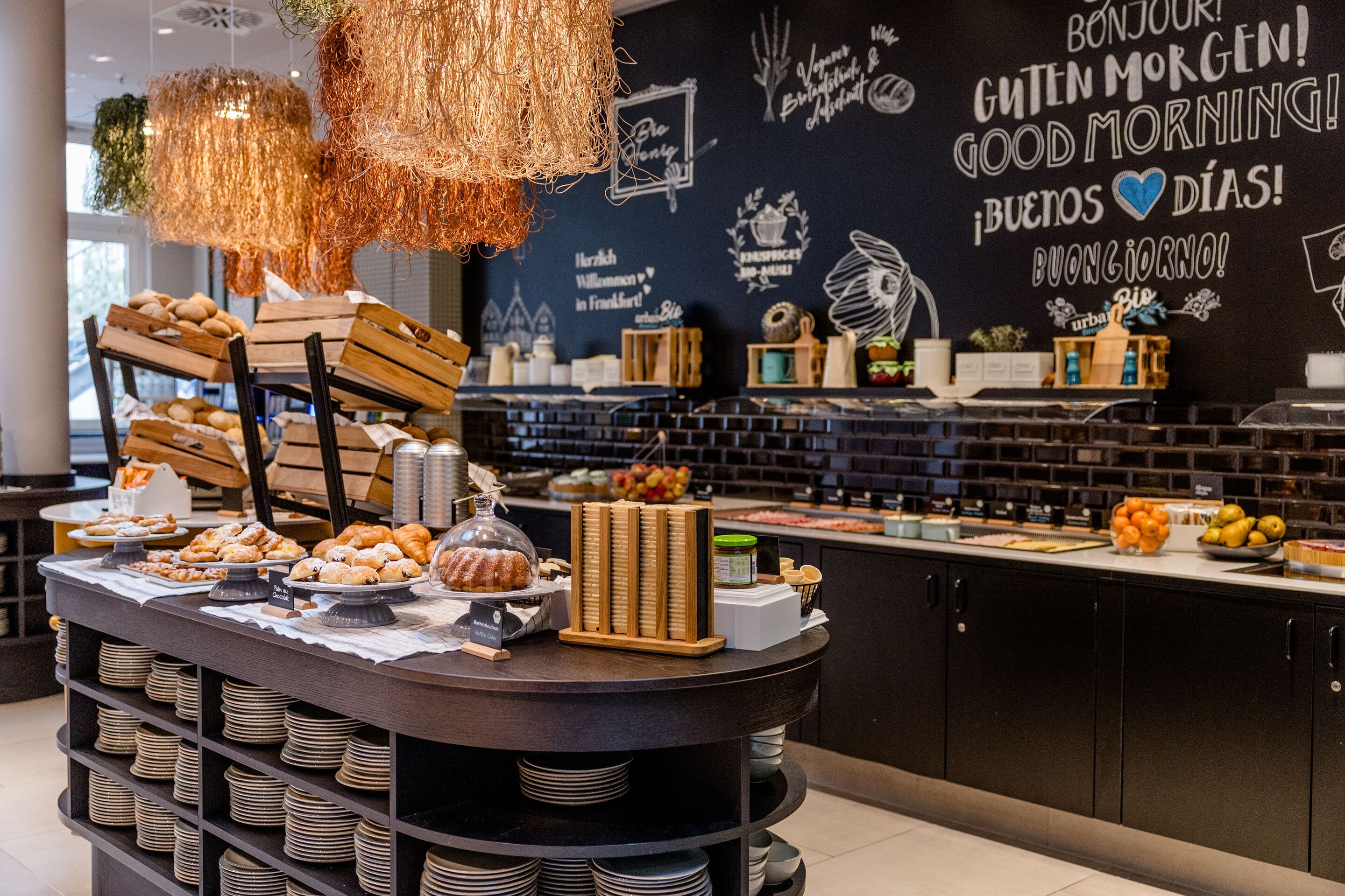 Hotel Frankfurt-Hauptbahnhof Motel One Frühstücksbuffet mit Brot, Gebäck, Obst und Müsli in modernem, einladendem Ambiente mit freundlicher Wandgestaltung und warmem Licht.