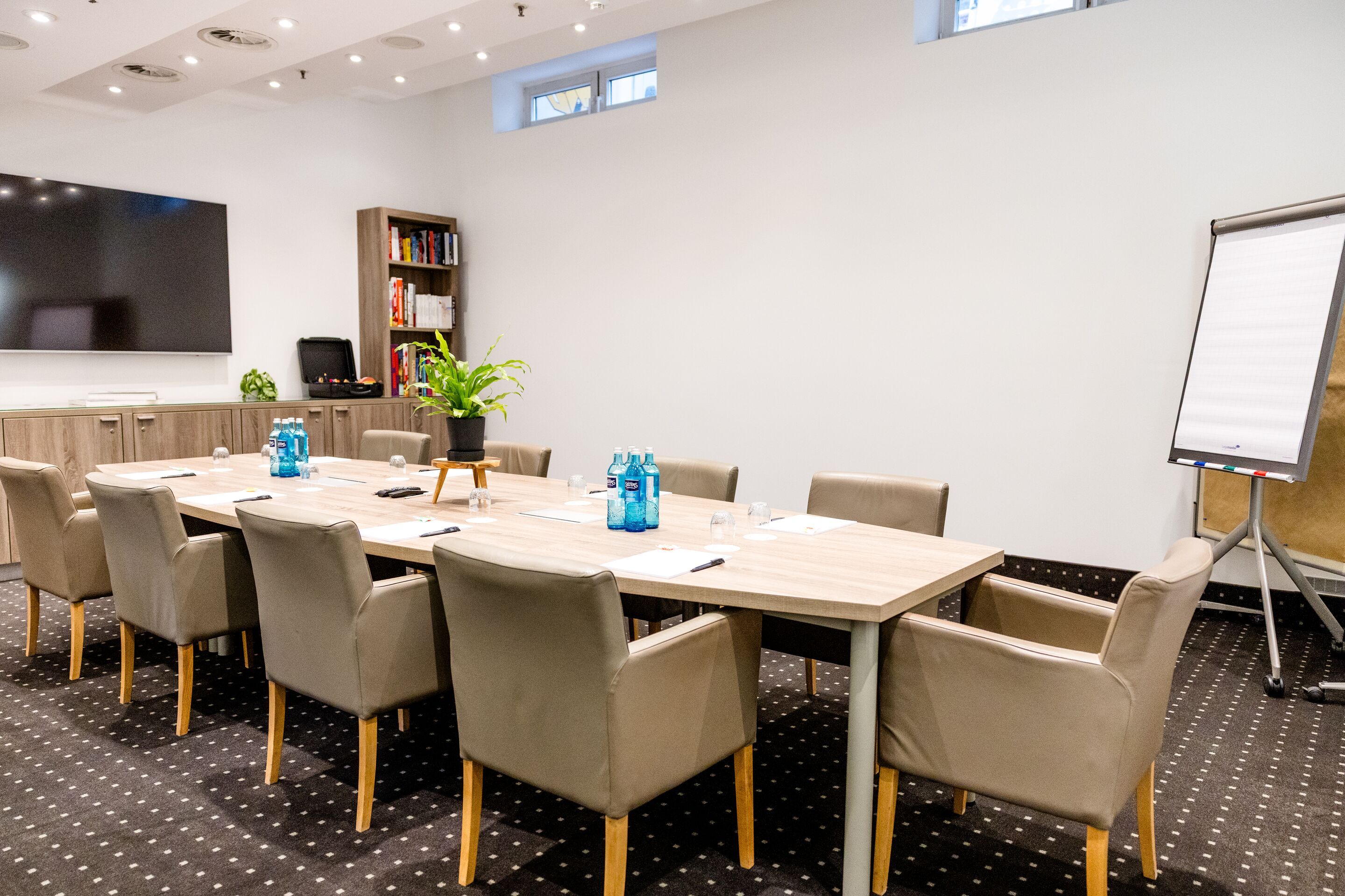 Hotel Frankfurt-Hauptbahnhof Motel One Konferenzraum mit großem Holztisch, Polstersesseln, Flipchart und Fernseher, ideal für Meetings und Präsentationen in heller Atmosphäre.