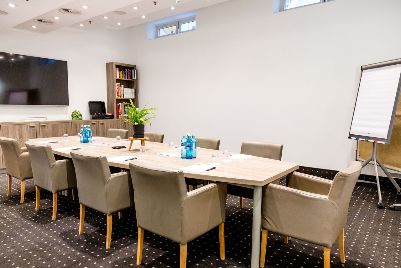 Hotel Frankfurt-Hauptbahnhof Motel One Konferenzraum mit großem Holztisch, Polstersesseln, Flipchart und Fernseher, ideal für Meetings und Präsentationen in heller Atmosphäre.
