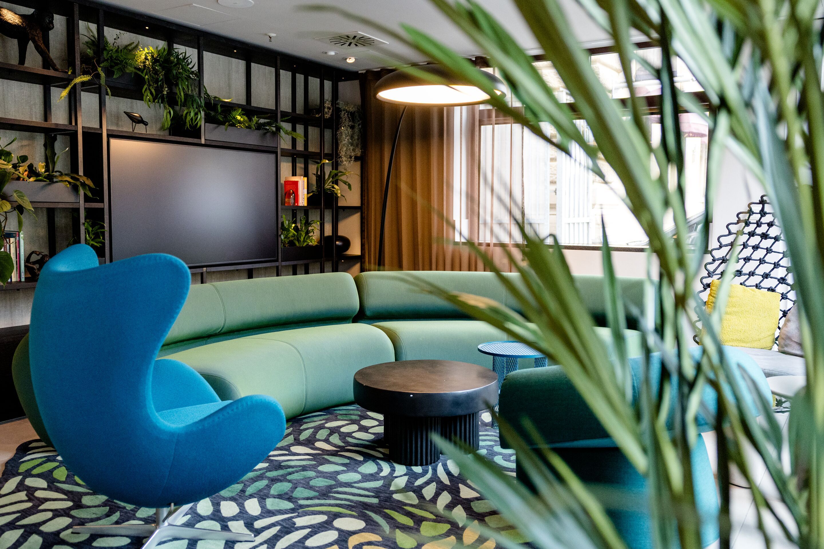 Hotel Frankfurt-Hauptbahnhof Motel One Lounge mit grüner, geschwungener Couch, Design-Sessel und rundem Couchtisch, umgeben von Pflanzen und natürlichem Licht.
