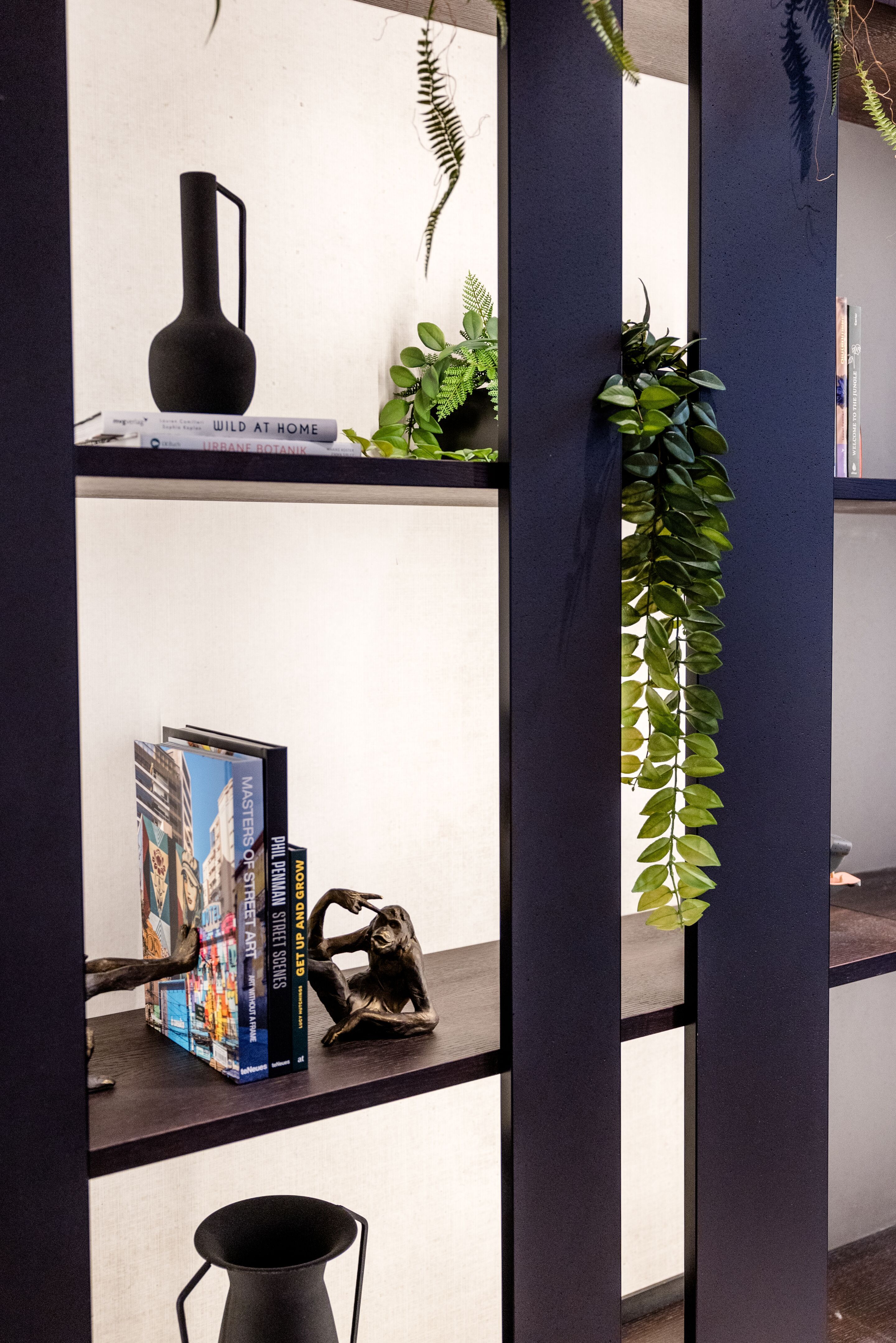 Designhotel Frankfurt-Hauptbahnhof Motel One Wandregal mit Dekorationen wie eine schwarze Vase, Bücher, eine Skulptur sowie grüne Pflanzen.
