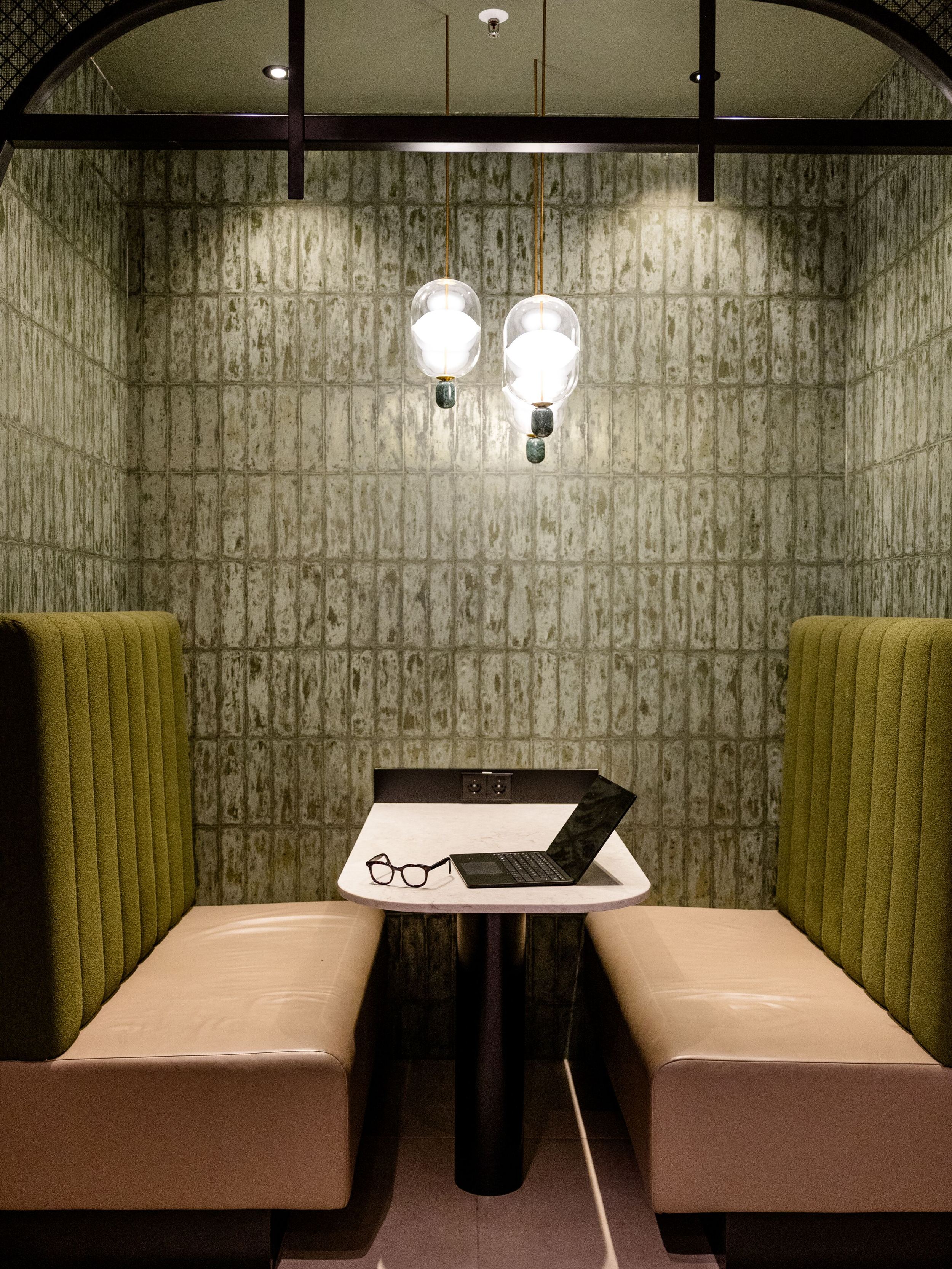 Hotel Frankfurt-Hauptbahnhof Motel One Sitzecke mit zwei gepolsterten Bänken und einem Tisch, beleuchtet von zwei Wandlampen. Ein Laptop und eine Brille liegen auf dem Tisch.