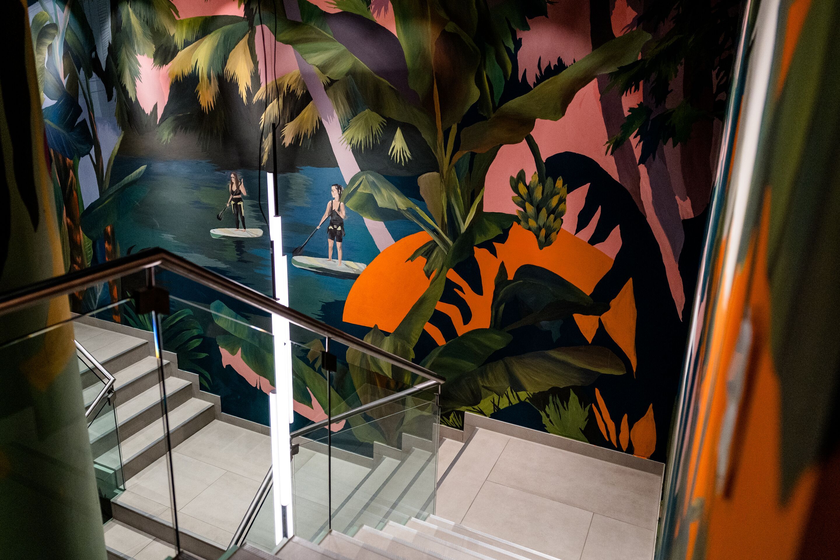 Hotel Frankfurt-Hauptbahnhof Motel One Treppenhaus mit beleuchtetem Glasgeländer. Wand hinter der Treppe zeigt farbenfrohes Tropen-Motiv mit Palmen, Wasser, und Sonne für entspannte Urlaubsstimmung.