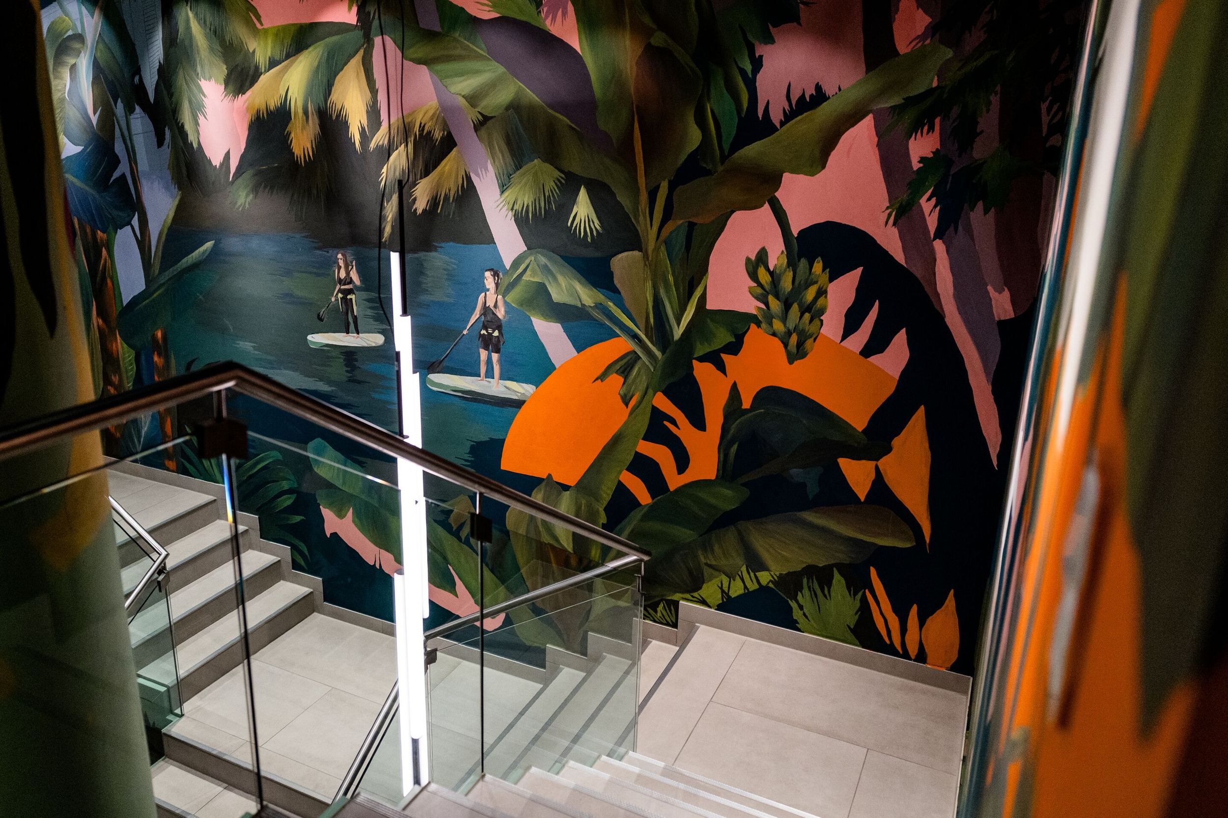 Hotel Frankfurt-Hauptbahnhof Motel One Treppenhaus mit beleuchtetem Glasgeländer. Wand hinter der Treppe zeigt farbenfrohes Tropen-Motiv mit Palmen, Wasser, und Sonne für entspannte Urlaubsstimmung.