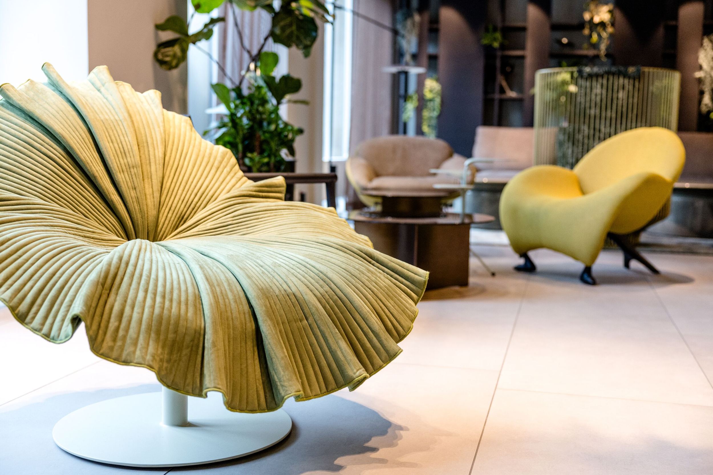 Hotel Frankfurt-Hauptbahnhof Motel One Lounge mit ausgefallenem, blattförmigem Sessel im Vordergrund und weiteren Sessels im Hintergrund, umgeben von Pflanzen und gemütlicher Atmosphäre.