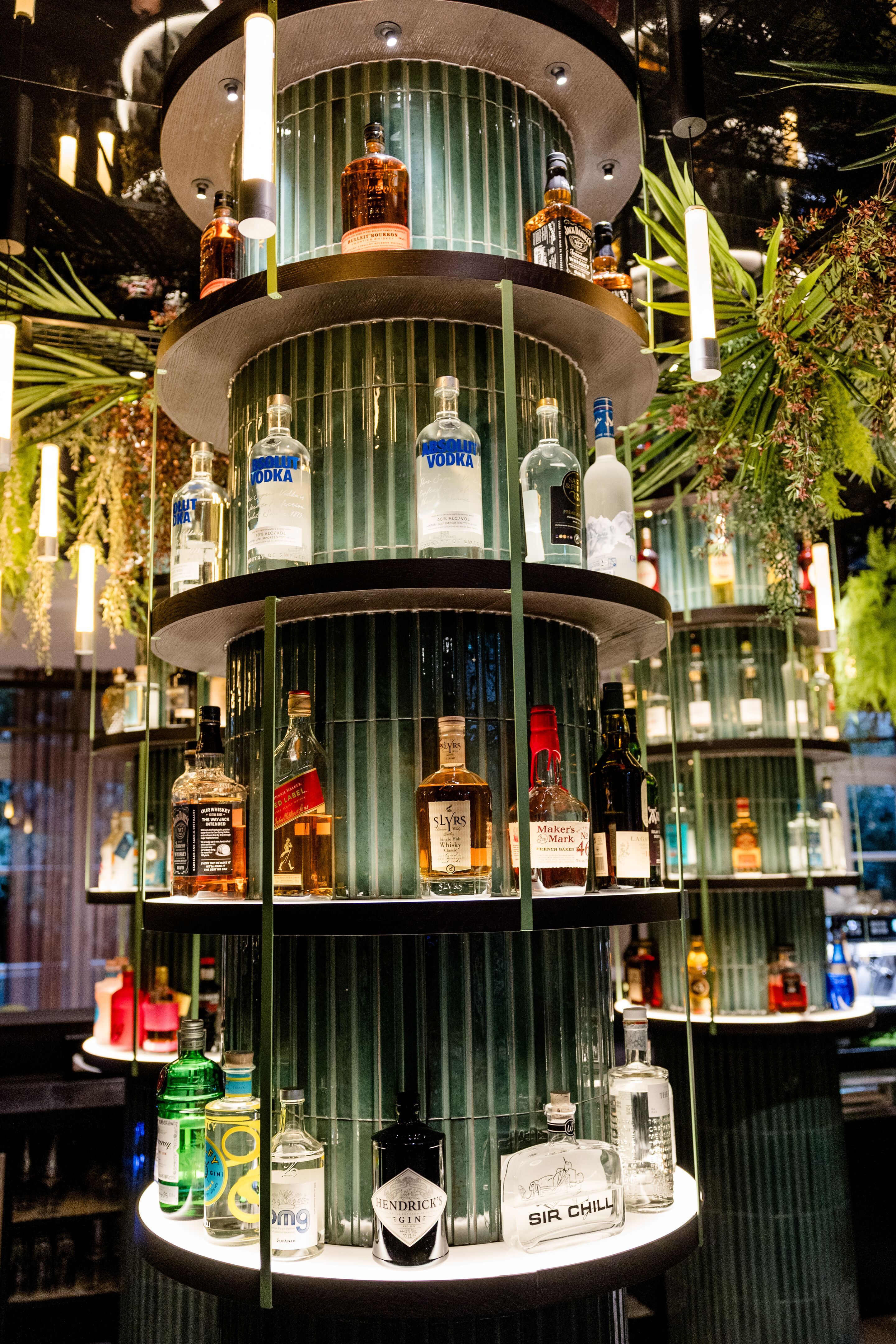 Hotel Frankfurt-Hauptbahnhof Motel One mehrstöckiges beleuchtetes Regal mit verschiedenen Spirituosenflaschen, umgeben von grünen Pflanzen, in stilvoller Bar-Atmosphäre.